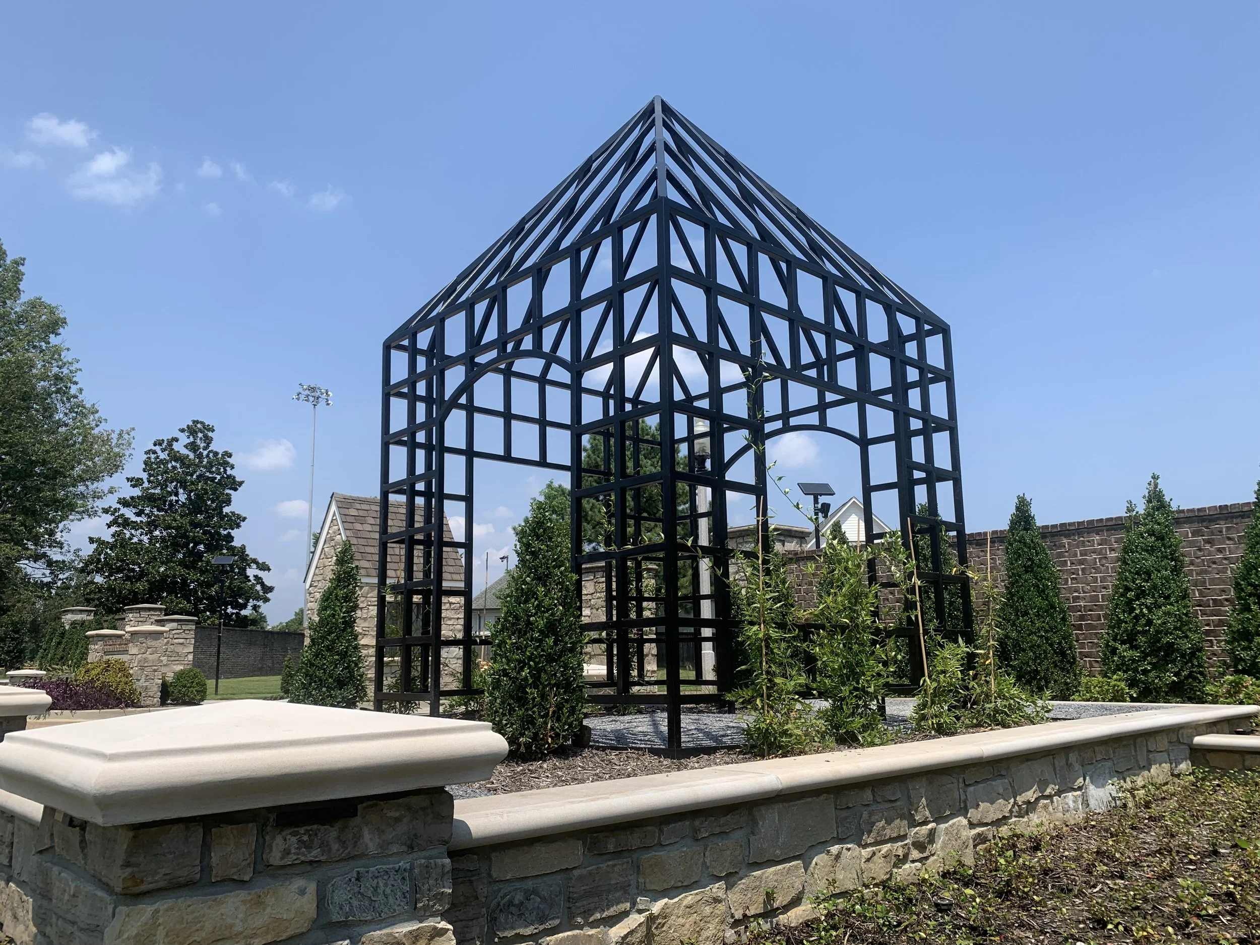 Metal garden trellis with arch and triangular roof shape, surrounded by small shrubs and plants, placed on a stone and concrete border in a landscaped yard.