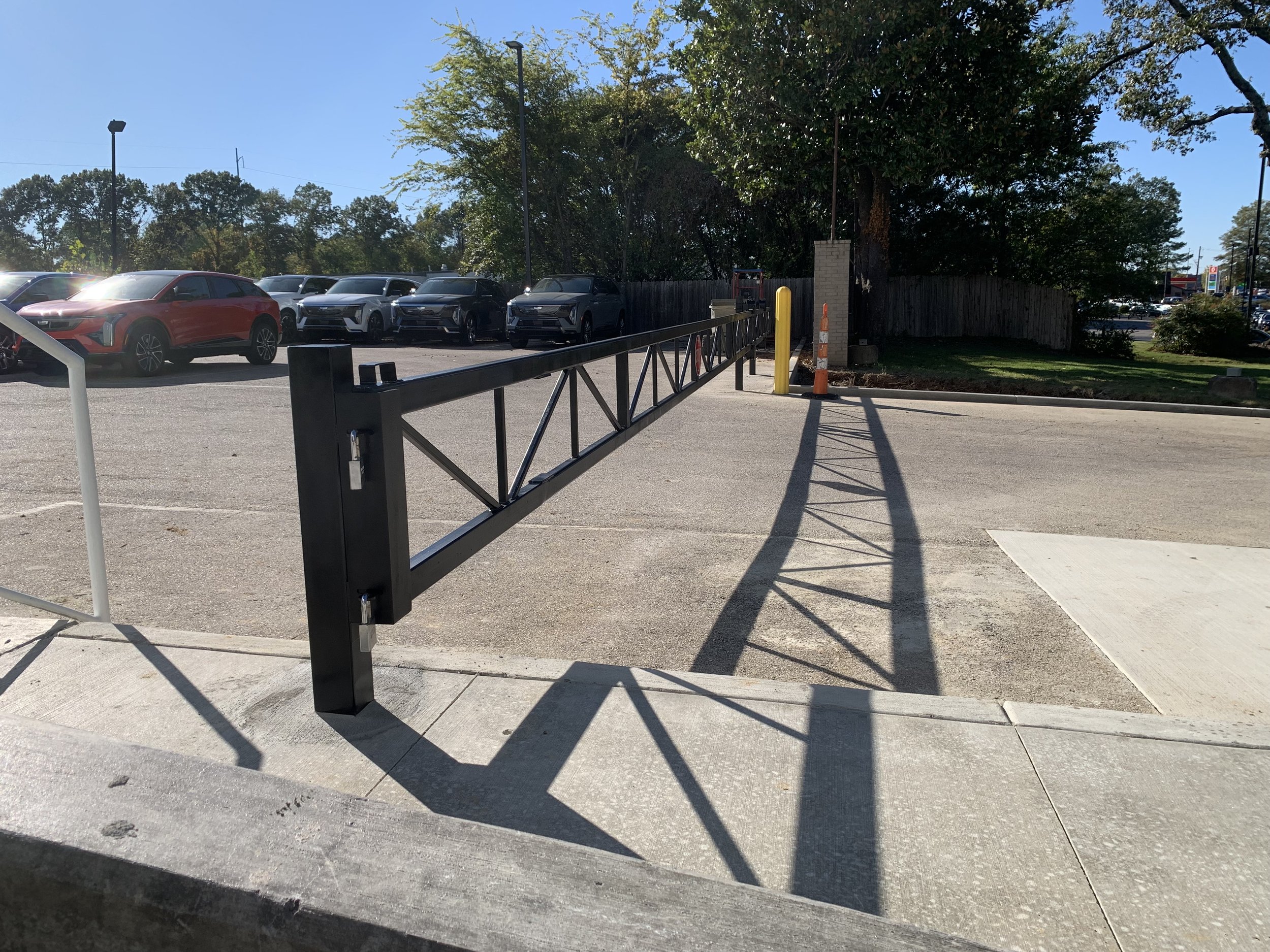 Parking lot with cars and a black metal security barrier, casting a shadow on the sidewalk, with trees and a blue sky in the background.