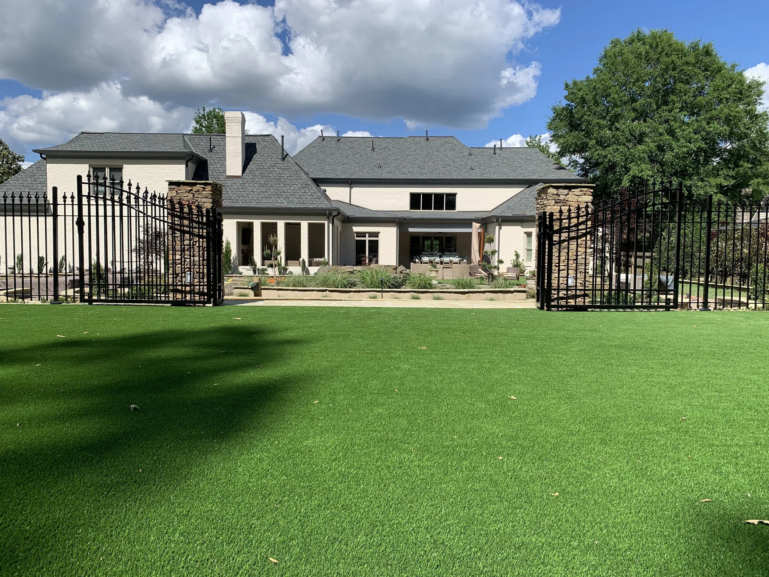 Backyard of a large white house with gray roof, gated entrance, green lawn, and landscaped garden with fencing, under cloudy sky.