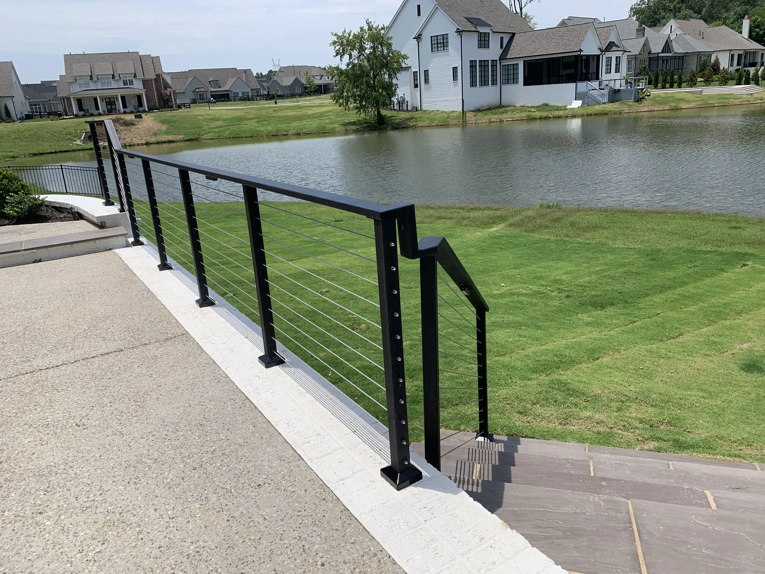 View of a residential area across a pond with houses and a well-maintained grassy lawn, taken from a balcony with black metal railing and concrete/tile flooring.