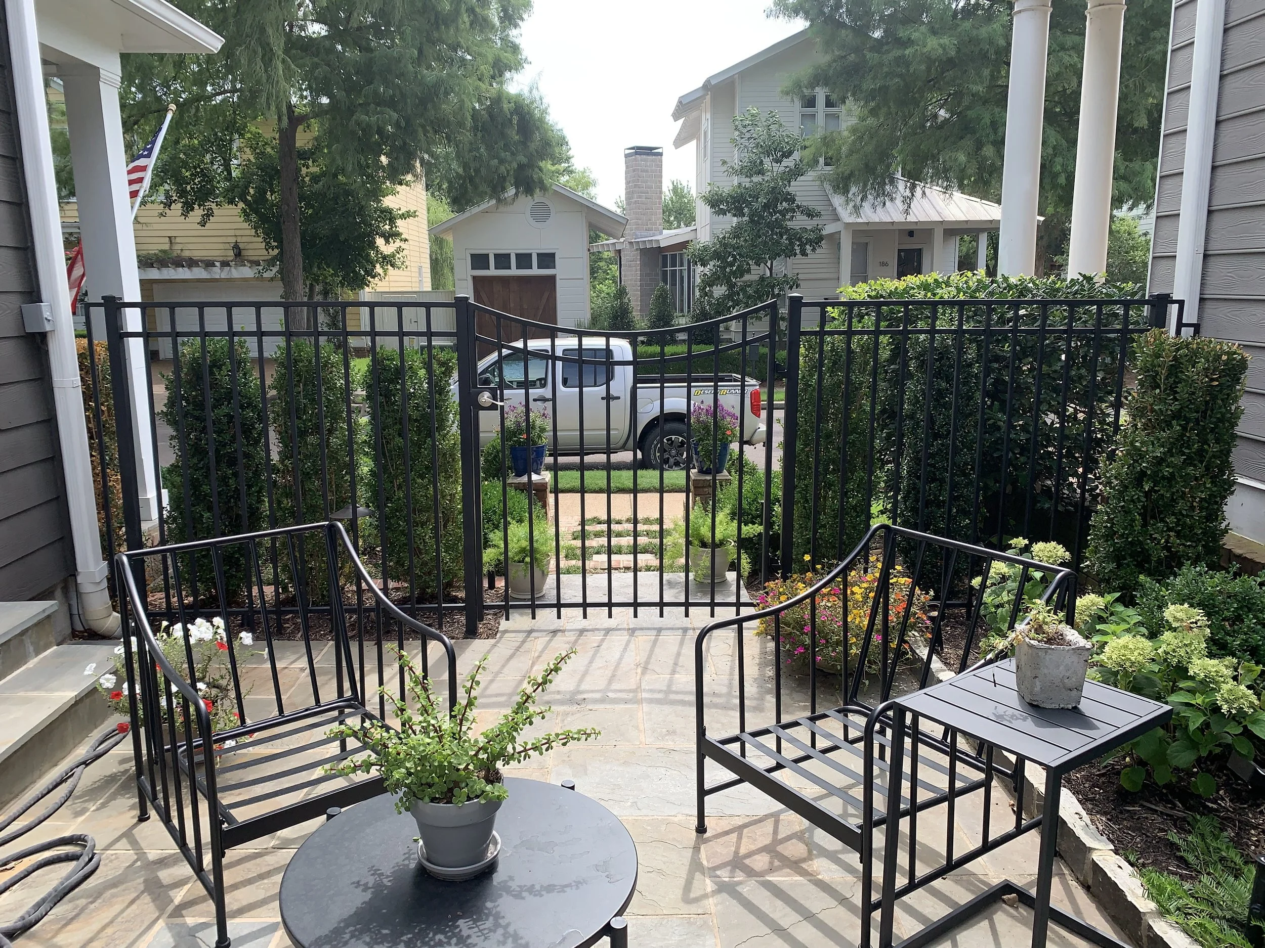 A small outdoor patio with black metal chairs, potted plants, and a garden area, enclosed by a black metal fence, with a view of a driveway and parked silver pickup truck in the background, and trees and houses around.