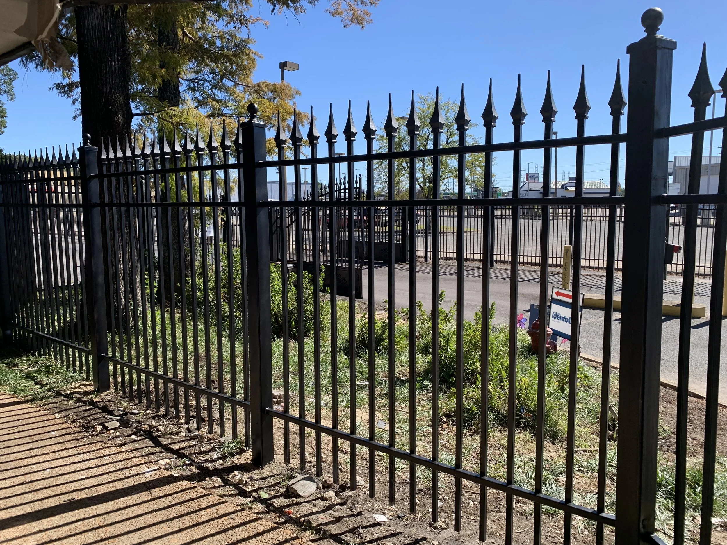 View of a black metal fence with pointed tips, with a small patch of greenery behind it and a parking lot with some vehicles in the background, under a clear blue sky.