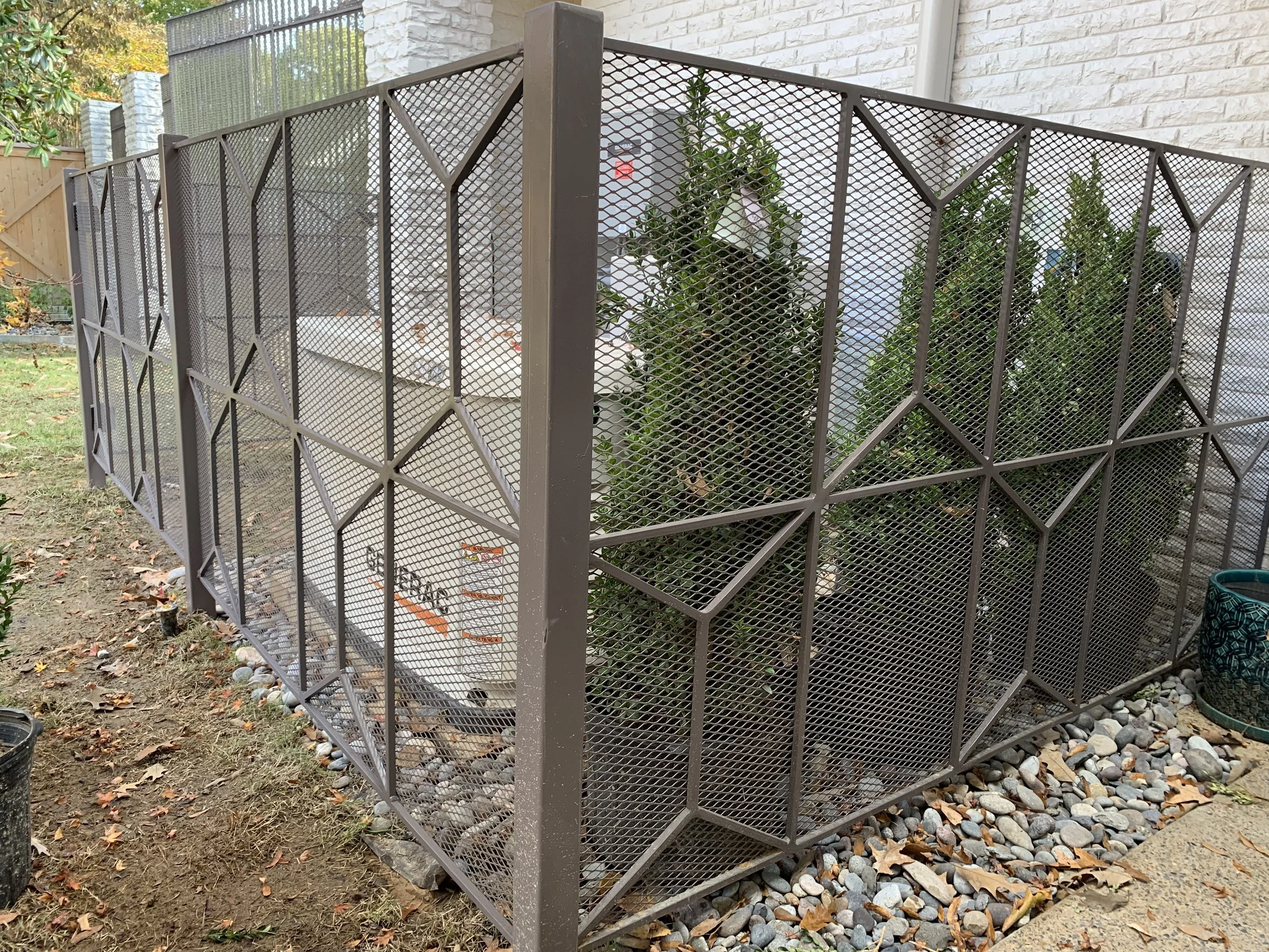 Small outdoor garden area enclosed by a metal lattice fence with rocks on the ground, plants, and a white building wall in the background.