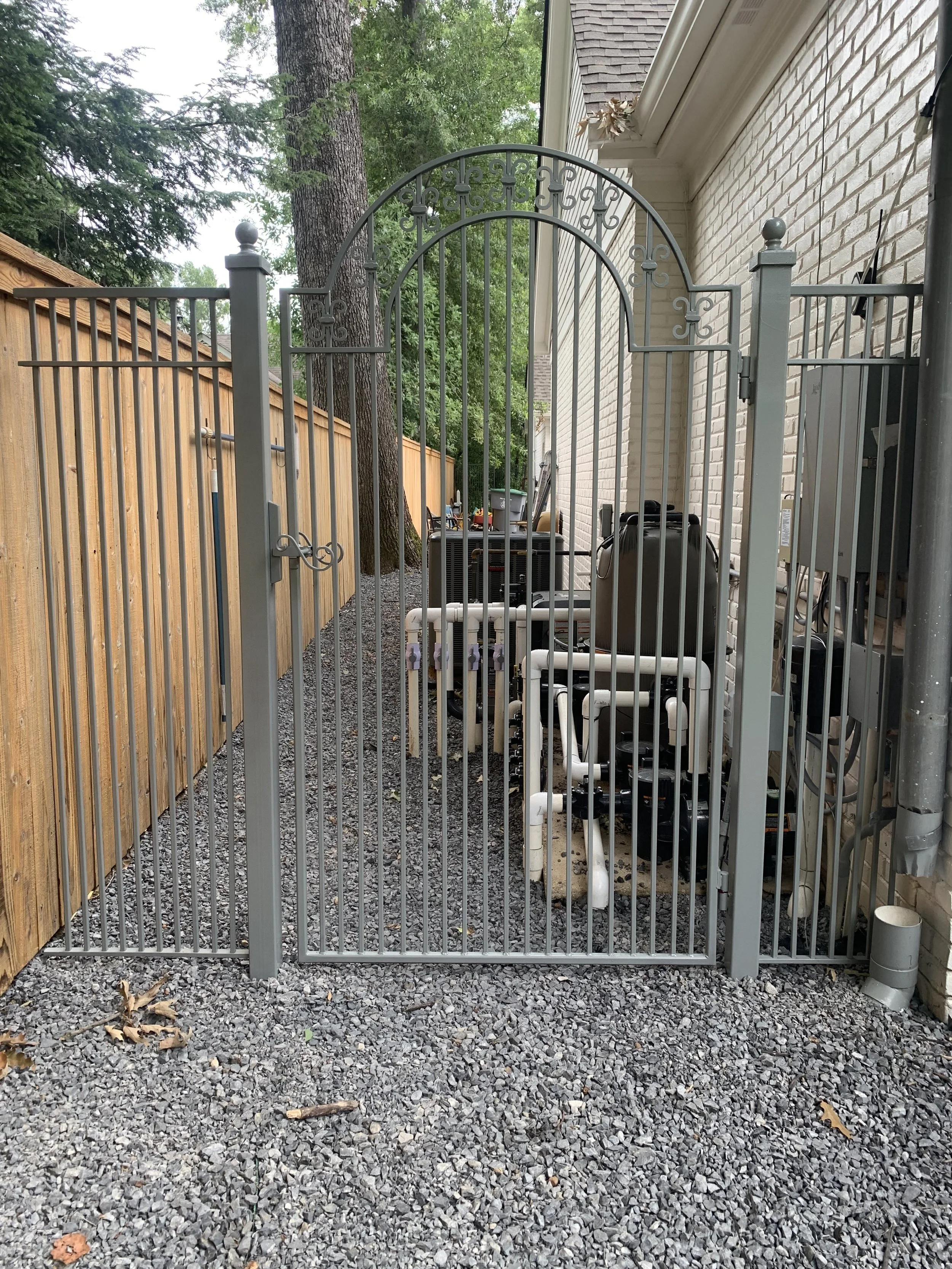 White metal gate opening to a small backyard enclosed by a wooden fence and brick house, with gravel ground, some tools and equipment, and tall trees in the background.