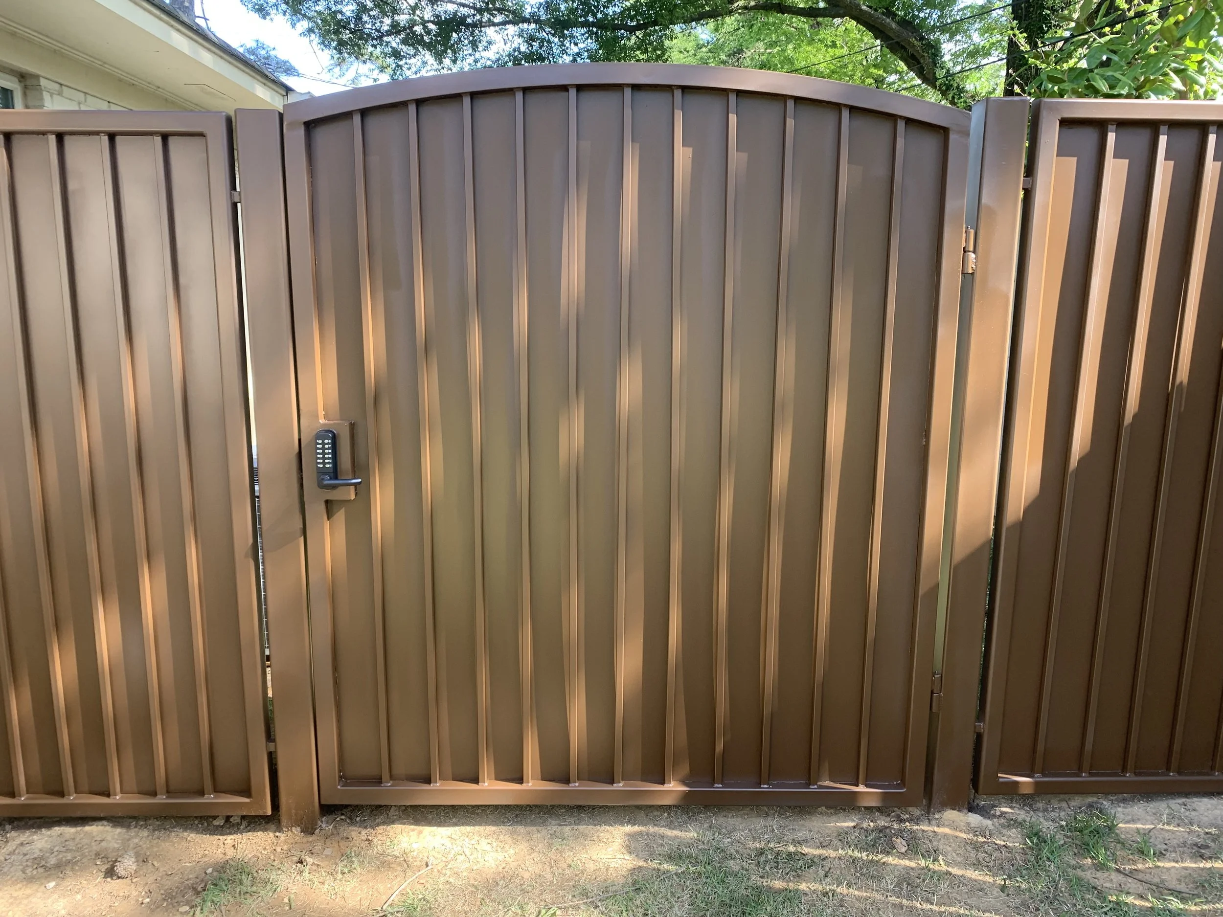 Brown metal gate with built-in keypad lock, partially open, leading to a backyard with trees and a house.