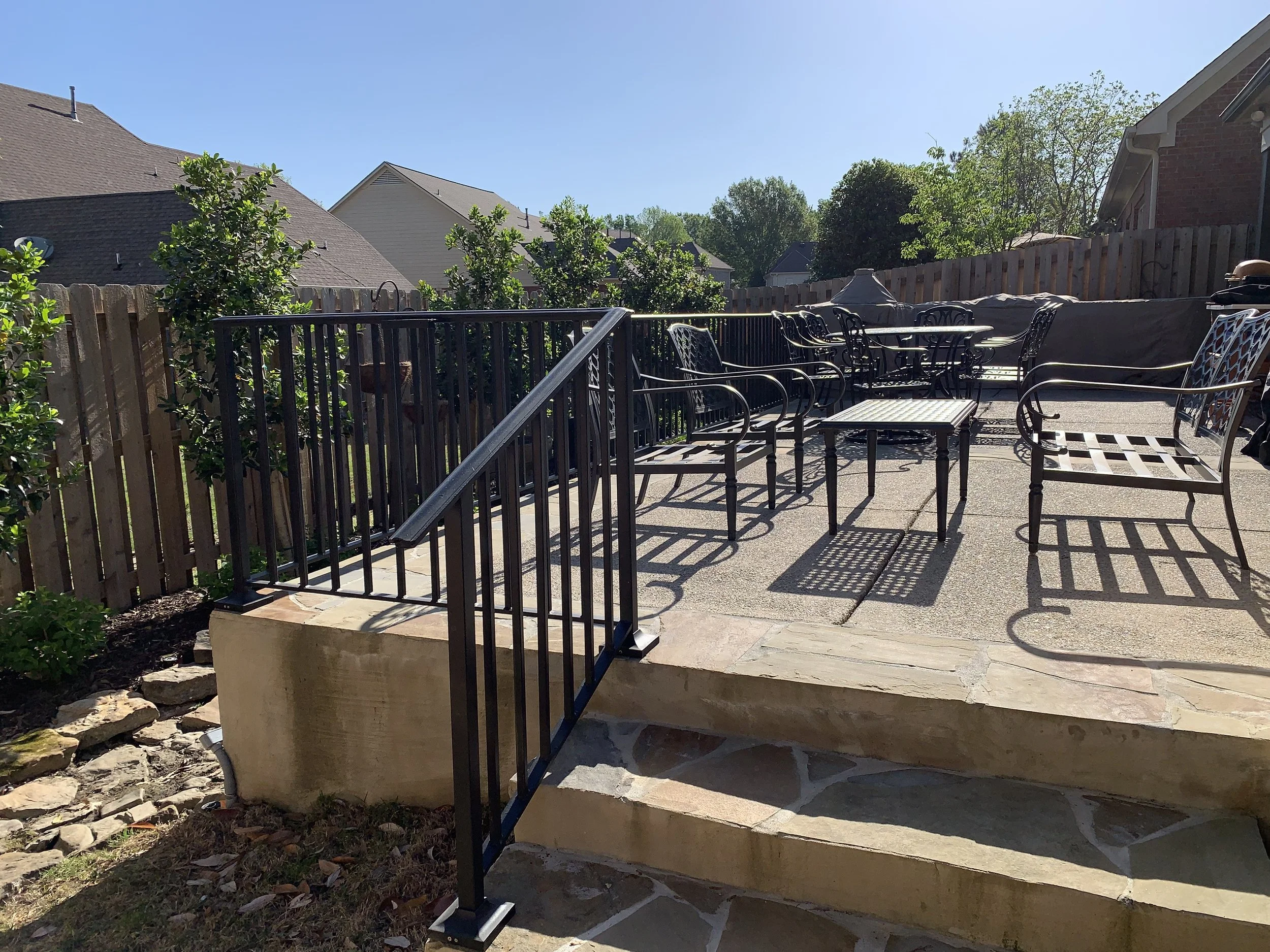 Backyard patio with outdoor furniture, stairs leading down, and a wooden fence with trees and houses in the background on a sunny day.