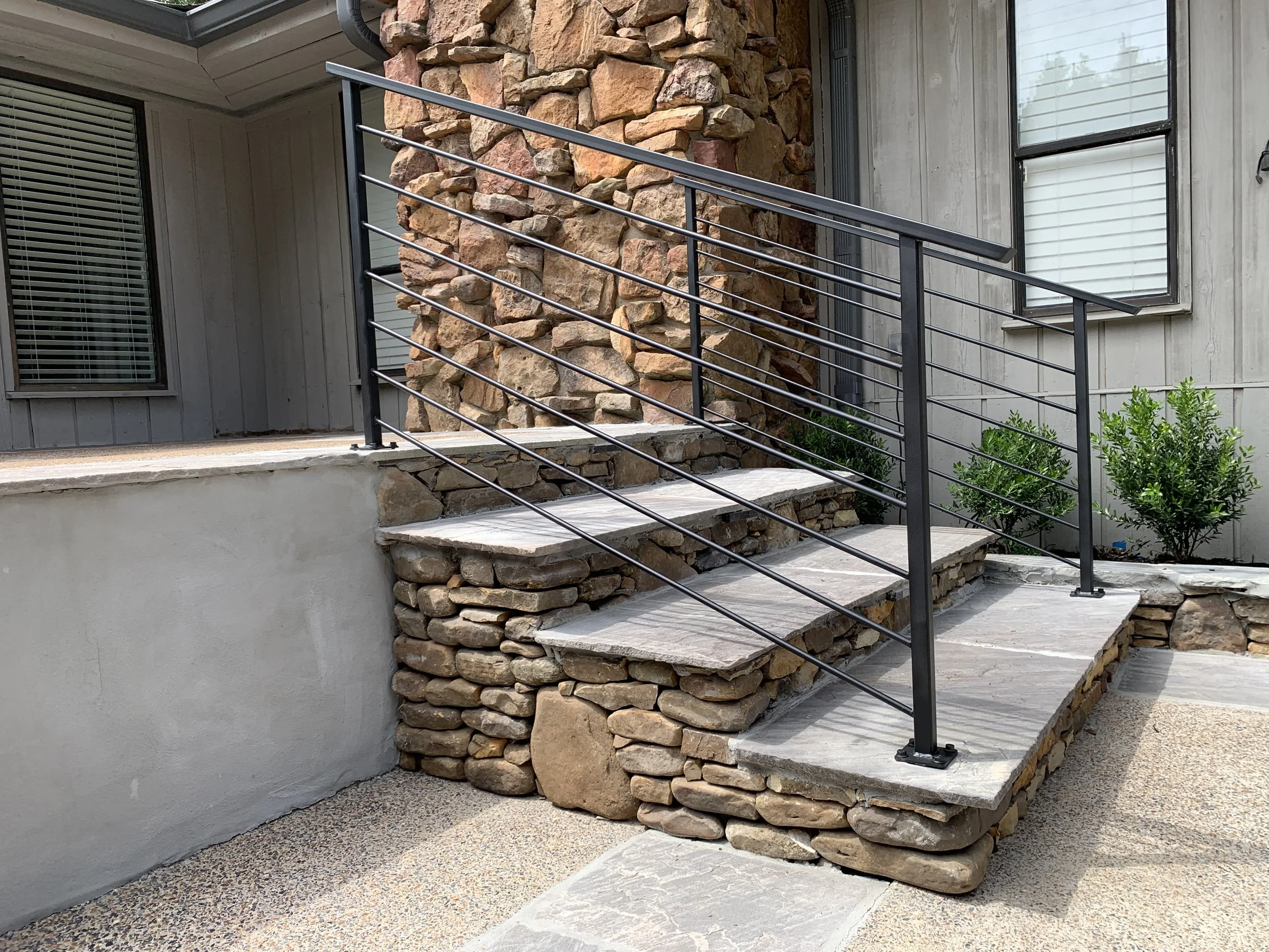 Outdoor concrete steps with a black metal railing leading up to a porch, with stone wall and bushes in the background.