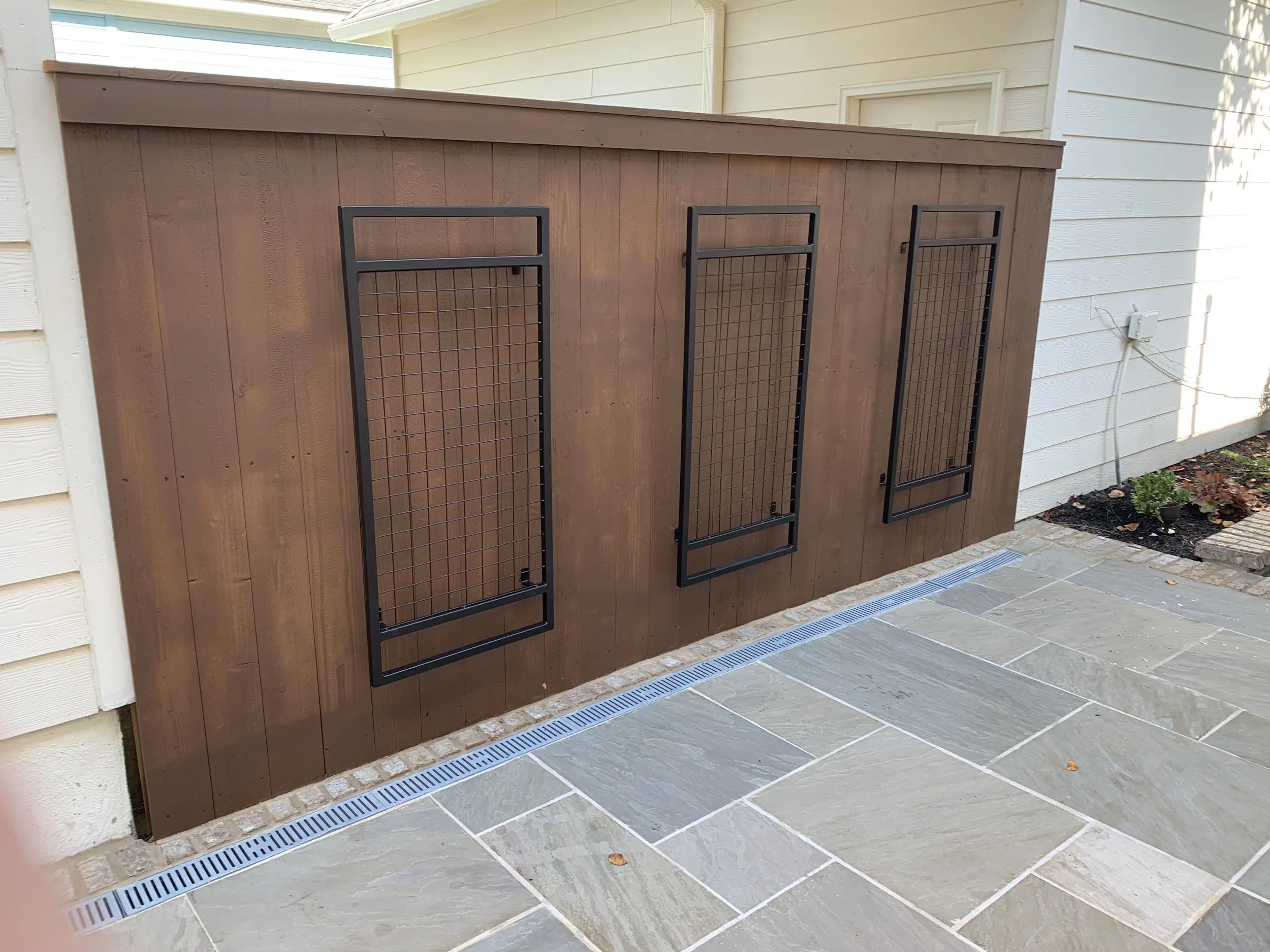 Three black metal pet doors installed in a wooden privacy fence, with a light-colored house wall and some small plants in the background.