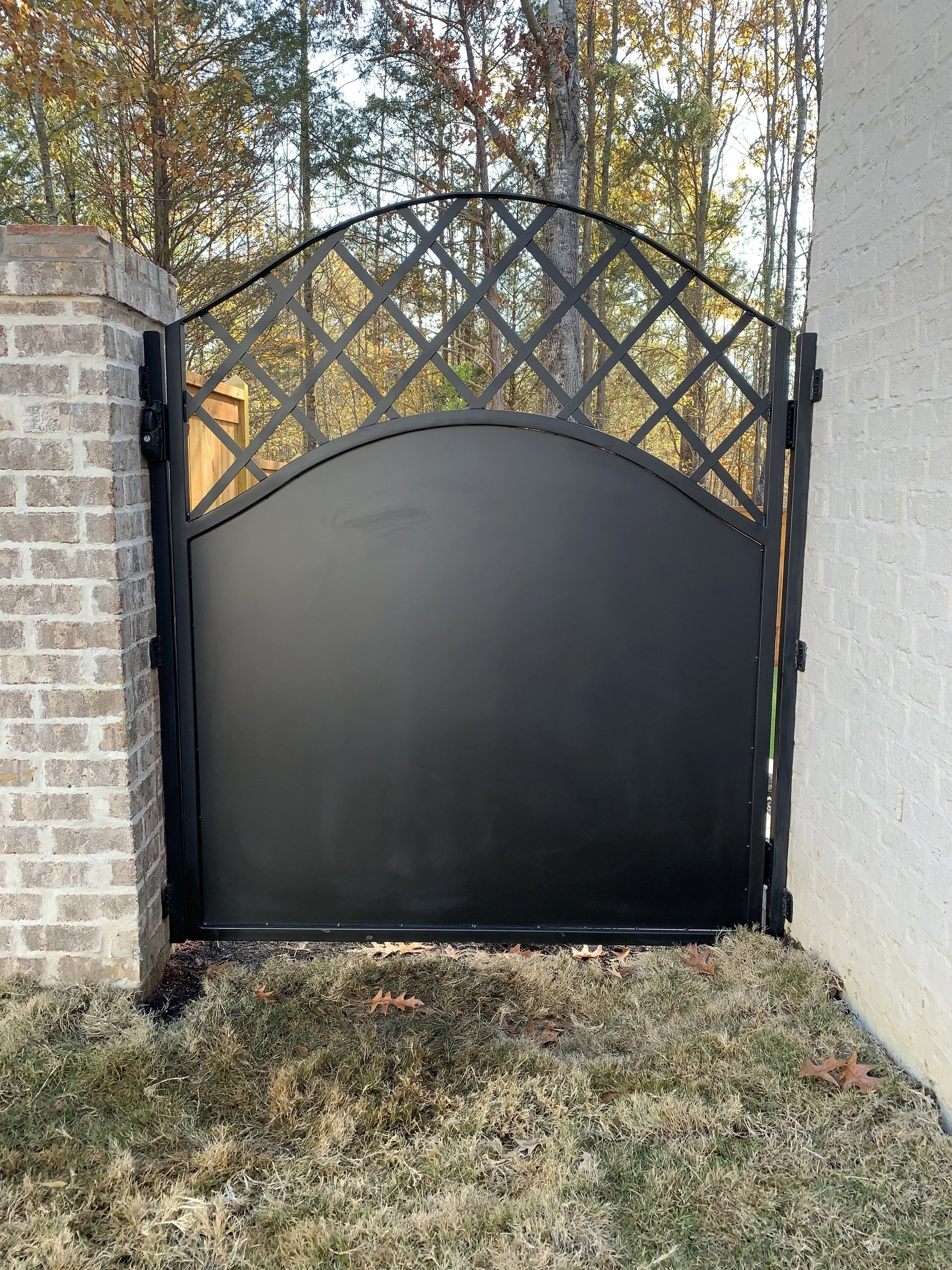 A black metal gate with a decorative lattice top, mounted between a brick pillar and a white brick wall, with trees and a wooden fence visible in the background.