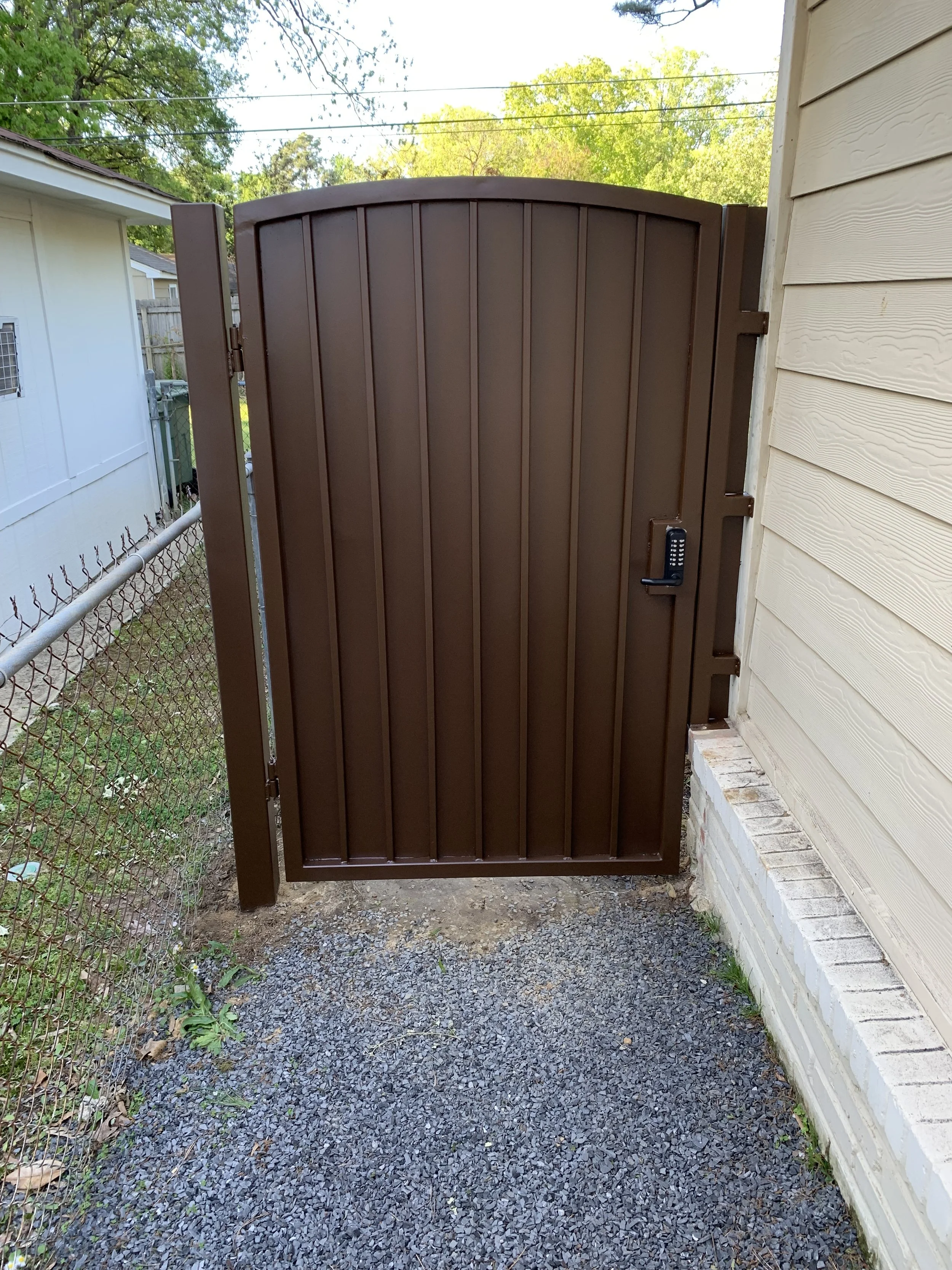 A brown metal gate with a keypad lock, situated next to a beige wooden house, on a gravel pathway with a chain-link fence on the left and green trees in the background.