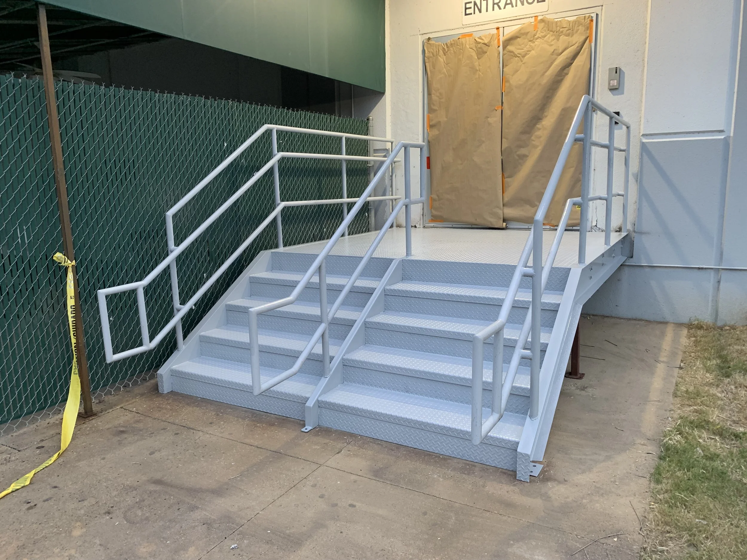 Metal wheelchair accessible ramp with rails leading up to a door covered by a brown paper sheet, with a sign above indicating 'ENTRANCE'.