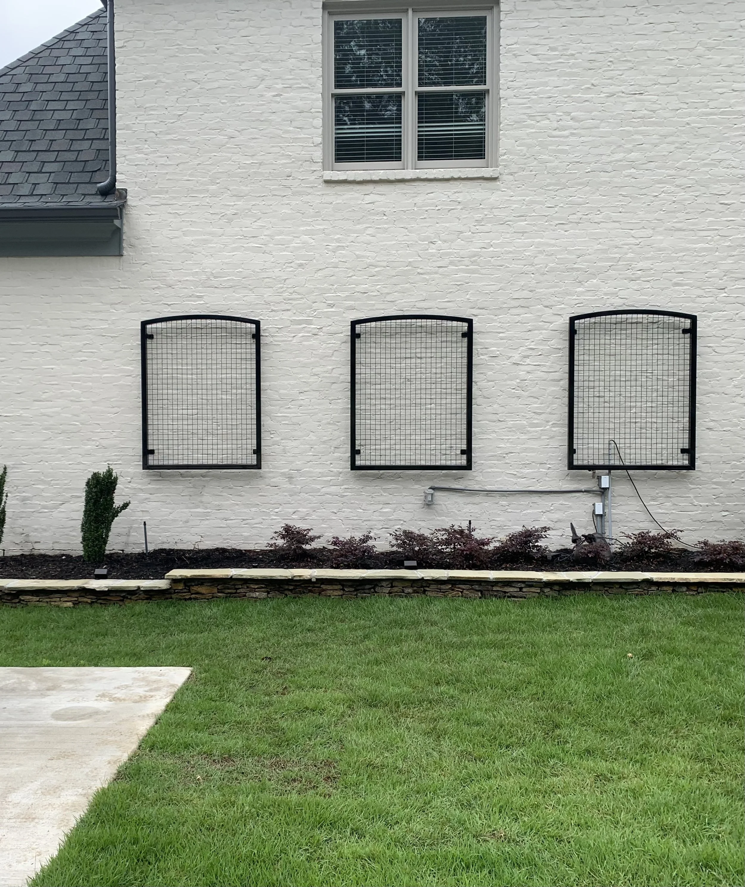 Three curved metal frames with wire mesh are mounted on a white brick wall, above a flower bed with purple plants, beside a window.