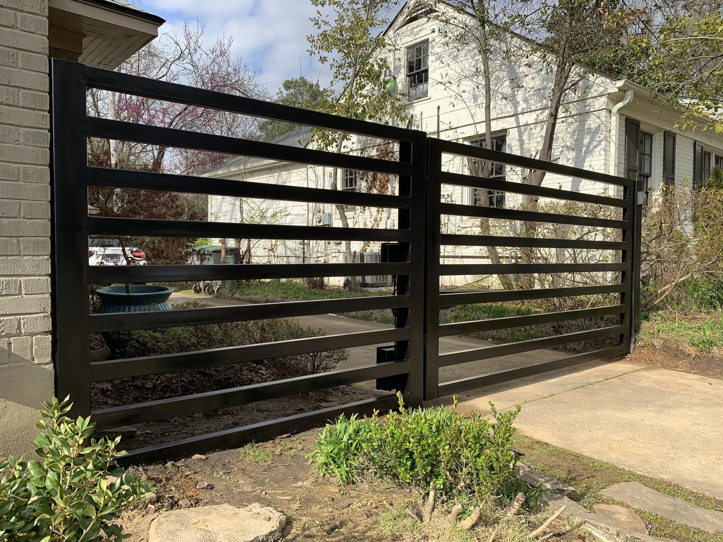 A black metal gate in front of a sidewalk and garden area, with a white house and trees in the background, under a blue sky with clouds.