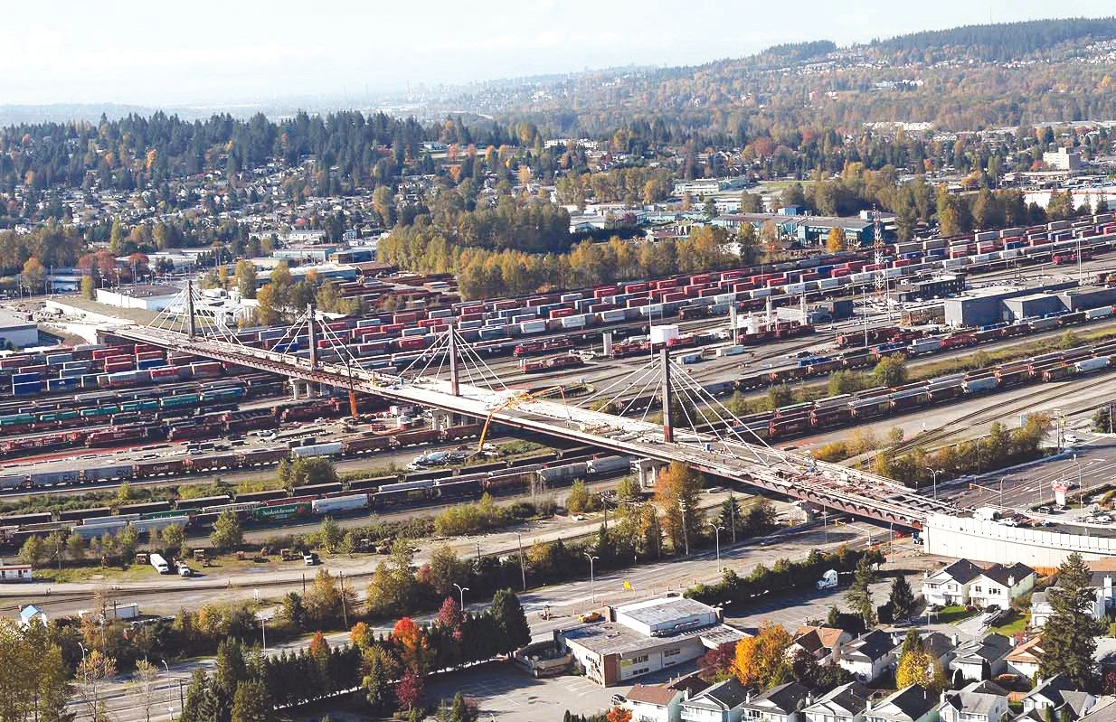 Aerial view of a large urban area with a prominent bridge that has cables and towers, surrounded by roads, train tracks, residential neighborhoods, and a hillside with trees in the background.