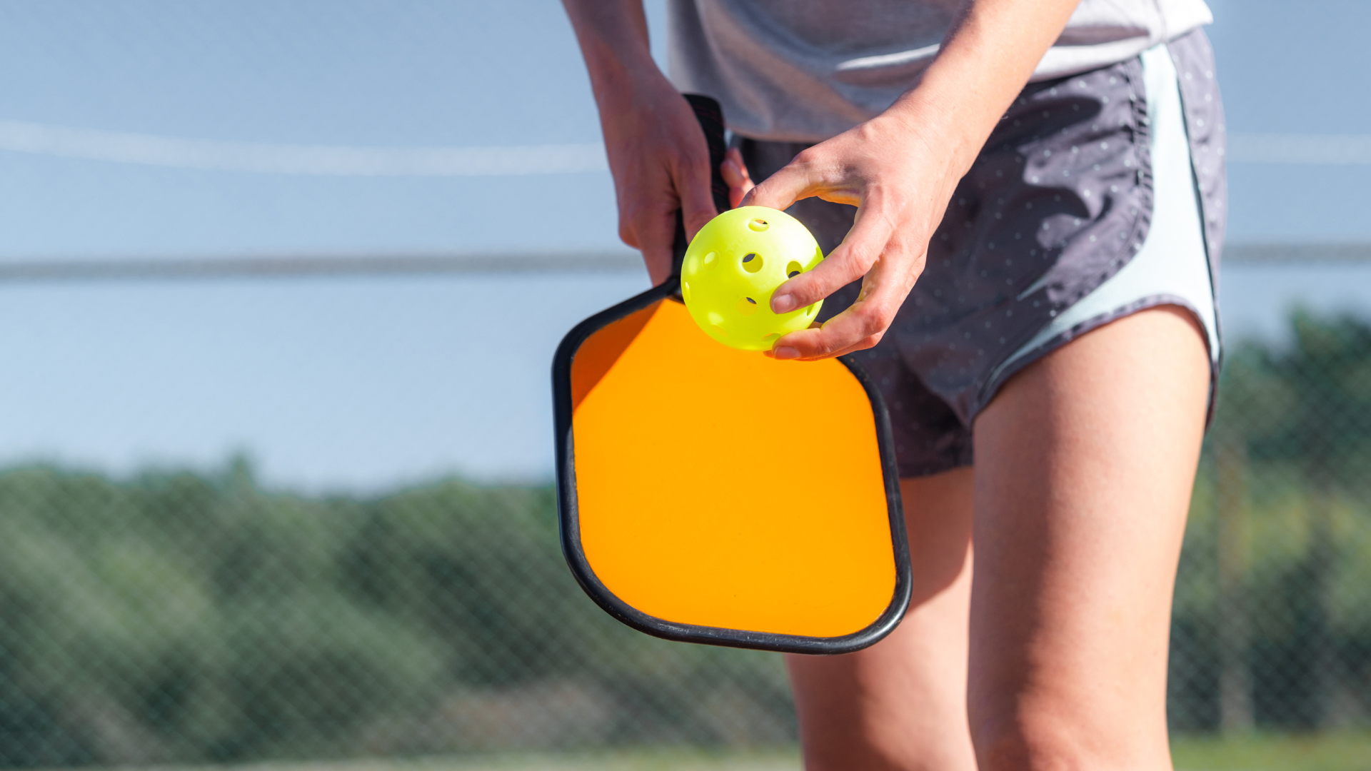 Close-up of a person playing pickleball holding a yellow perforated plastic ball with one hand, with a paddle and pickleball court in the background.