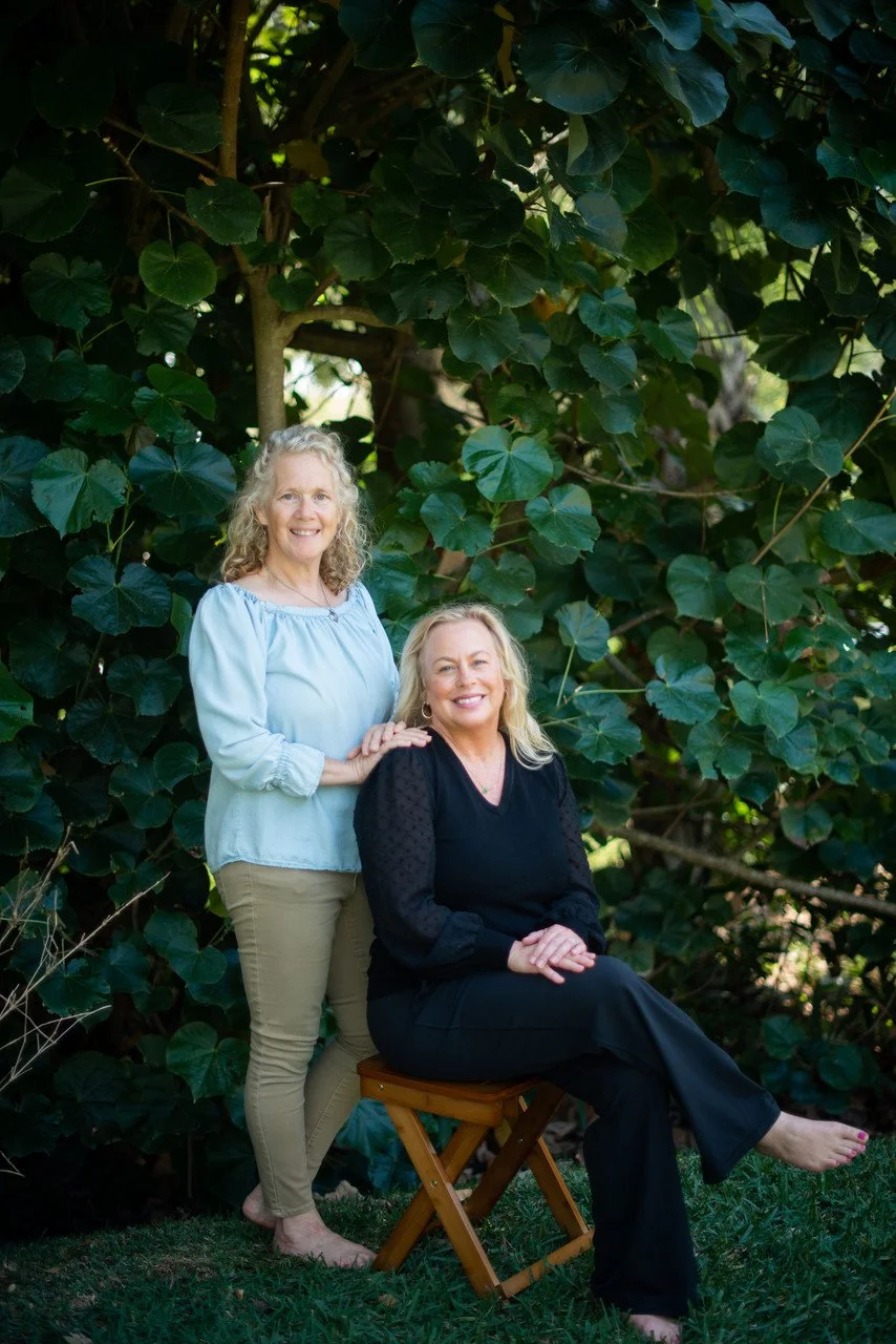 Two women, one standing and one seated on a small wooden stool, outdoors in front of lush green foliage, smiling at the camera.