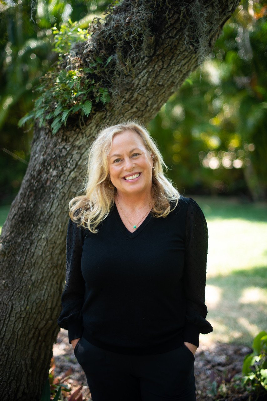 A smiling woman with blonde hair standing outdoors at the base of a tree, wearing a black long-sleeved top and black pants in a sunny, green park.