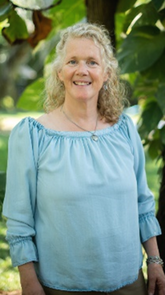 A woman with curly gray hair standing outdoors in front of lush green plants, smiling at the camera, wearing a blue blouse.