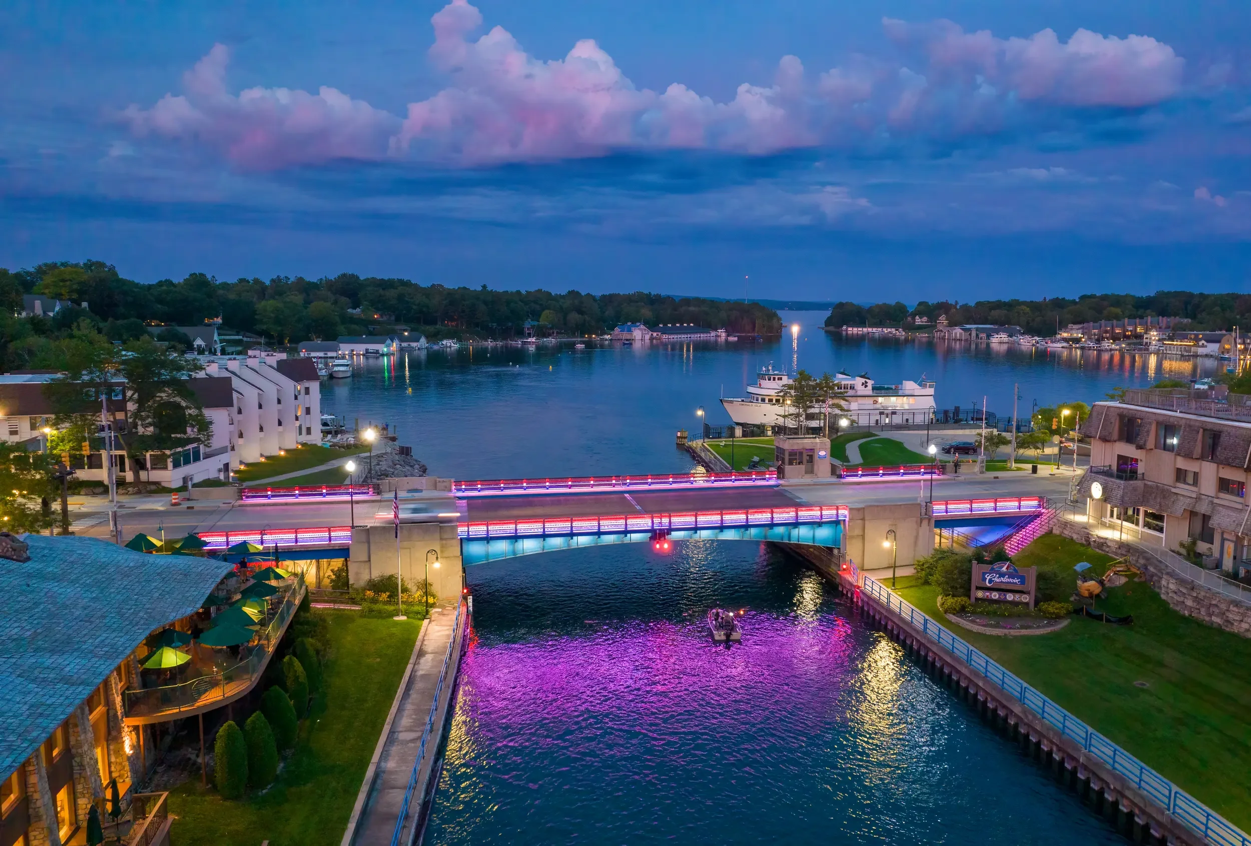 A picturesque evening view of a bridge over a canal with neon lights, surrounded by buildings, trees, and boats on the water under a cloudy sky.