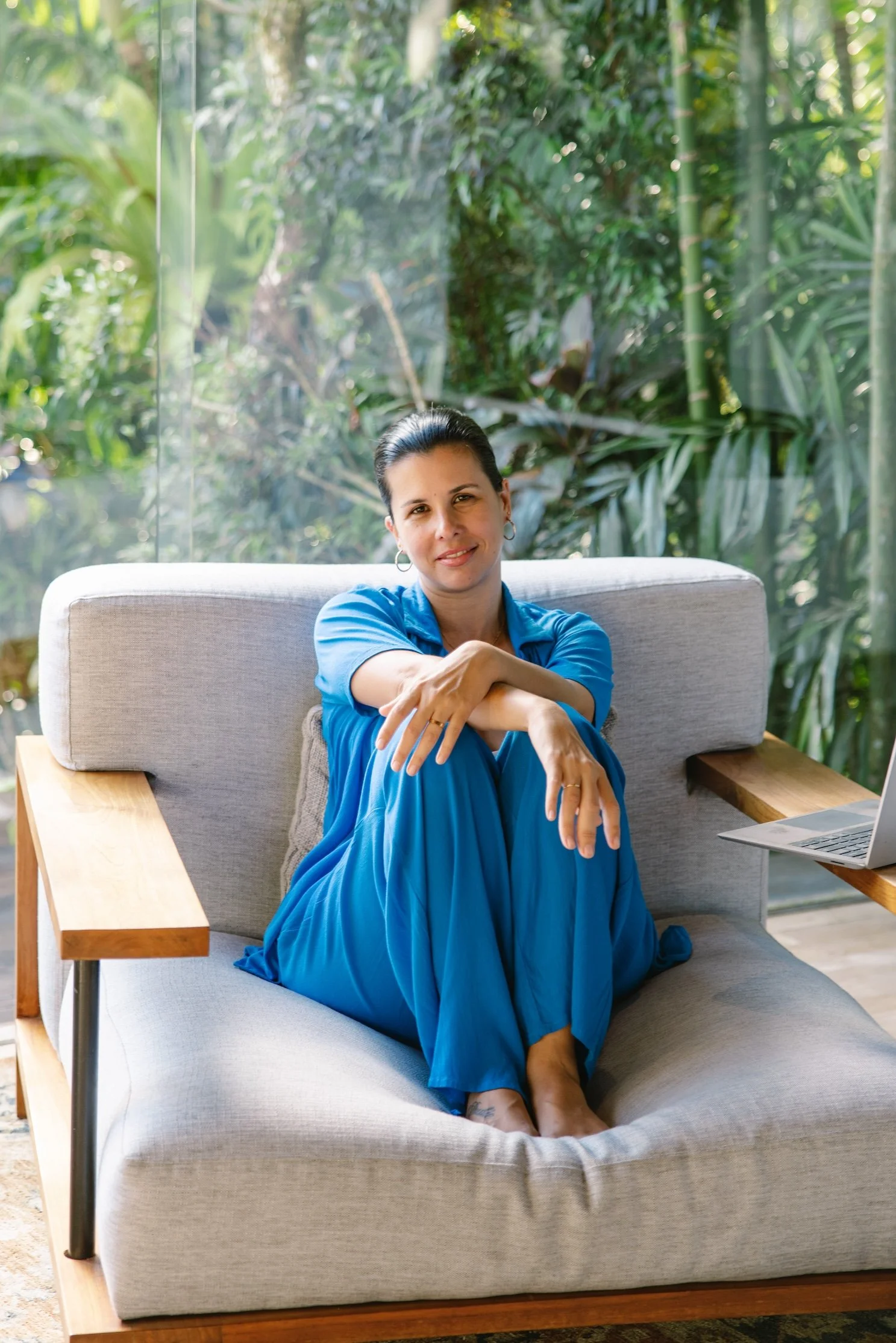 A woman with dark hair tied back, wearing a blue dress, sitting on a light-colored sofa. She has one arm across her knees and a gentle smile. The background features large windows with lush green plants outside.