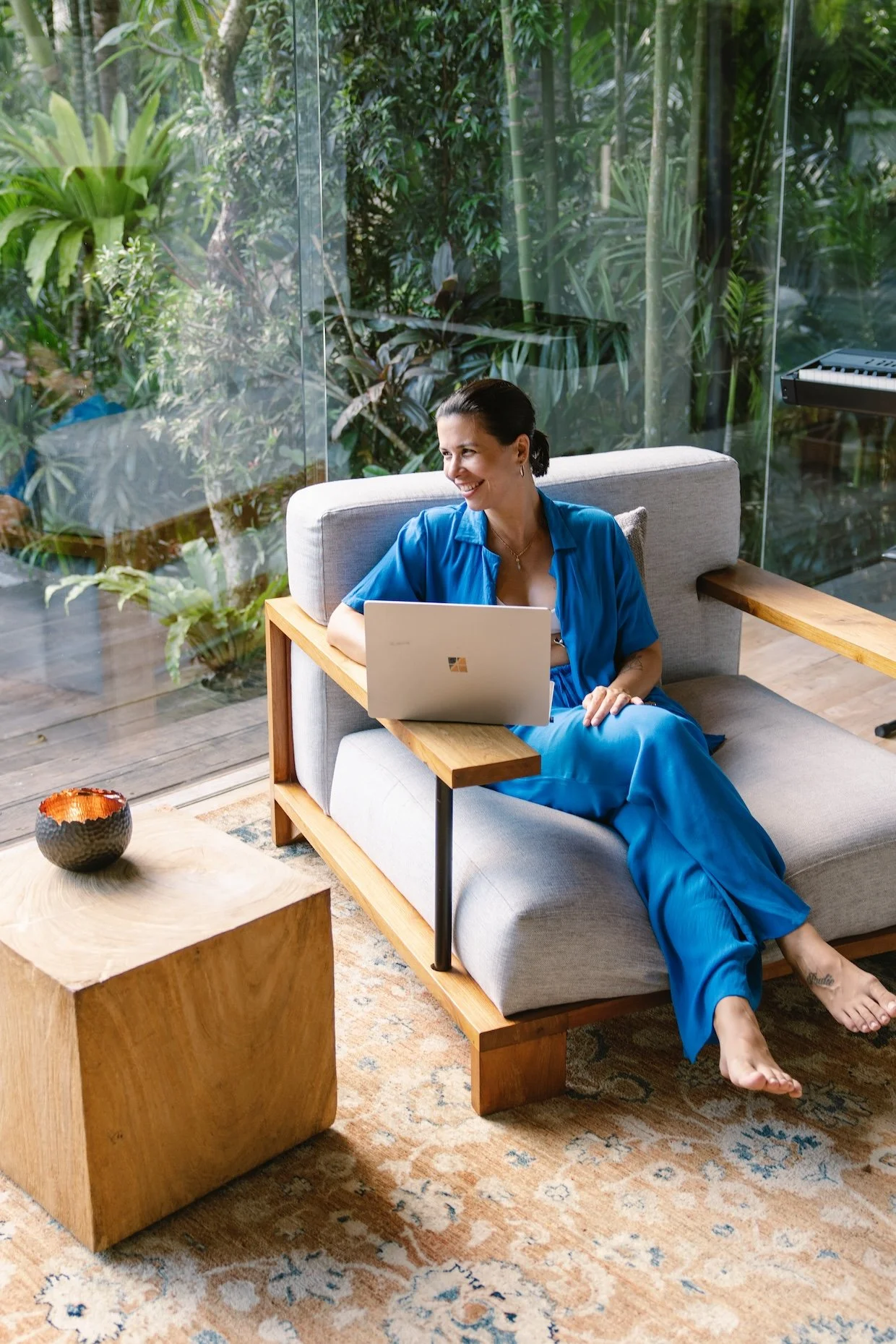 A woman sits on a modern gray sofa with wooden accents, working on a laptop. She is wearing a blue shirt and matching pants, smiling in a room with large glass windows and lush green foliage outside. There is a wooden side table with a decorative bowl and a patterned rug beneath.