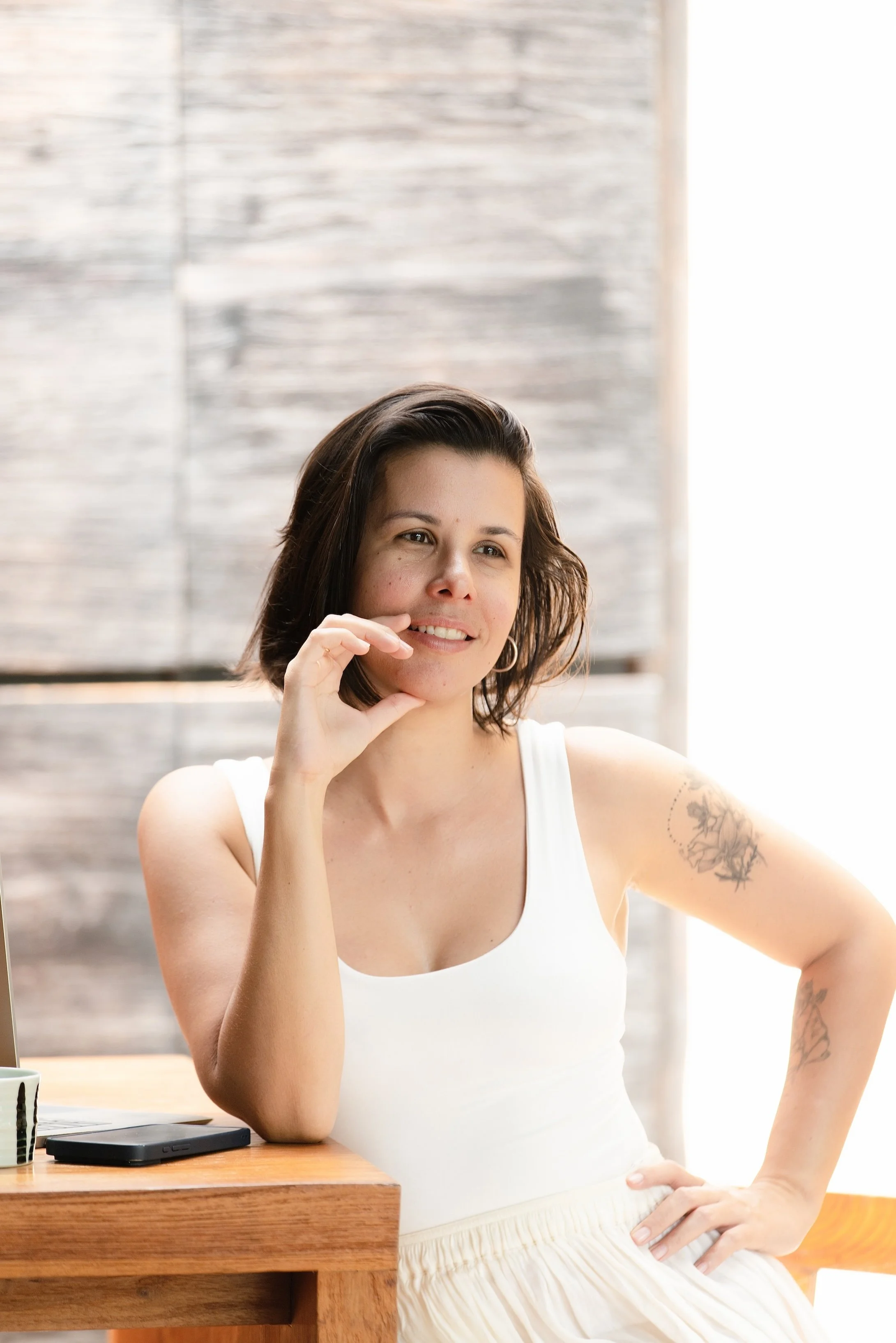 A woman with short brown hair, wearing a white tank top and cream-colored skirt, sitting at a wooden table with a smartphone and a cup nearby. She has tattoos on her right arm and is smiling softly, resting her chin on her right hand.