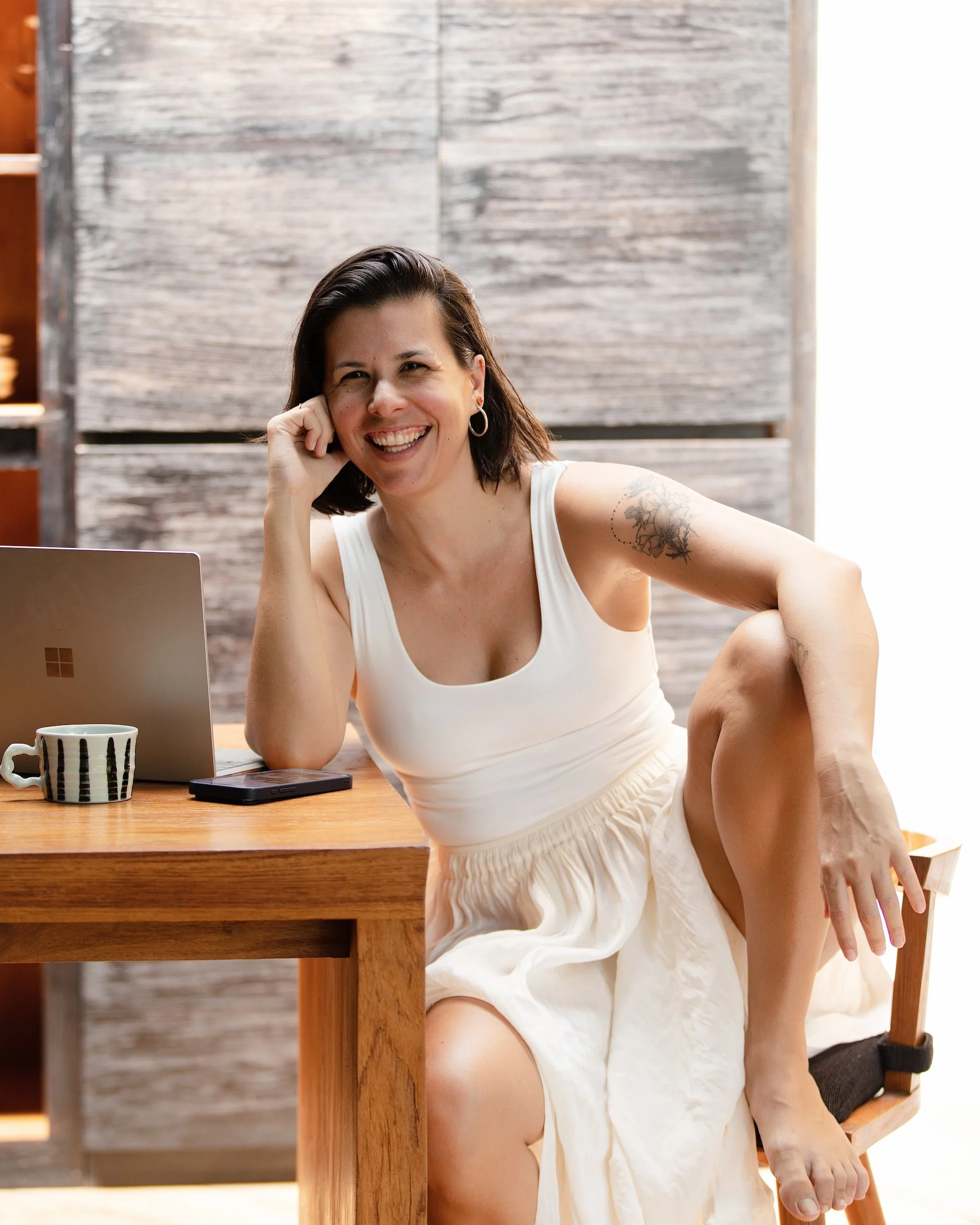 A woman with dark hair smiling and resting her head on her hand, sitting at a wooden table with a laptop, a coffee mug, and a smartphone in front of her, in a room with wooden paneling.