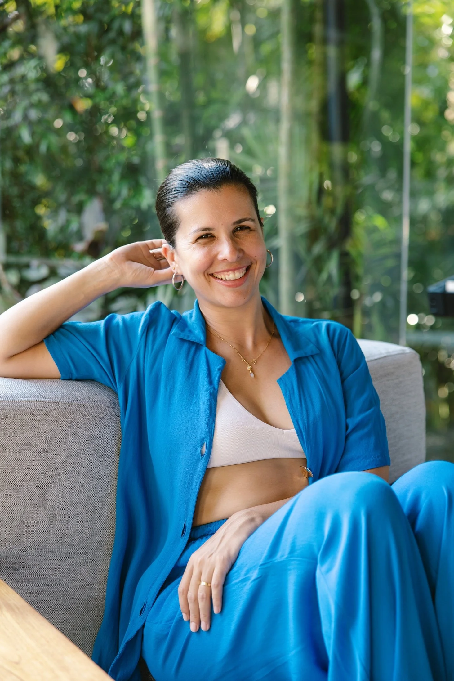 A woman smiling and relaxing on a sofa in a bright room with large green plants in the background. She is wearing a blue open shirt over a white top, with her hair slicked back, and gold hoop earrings.