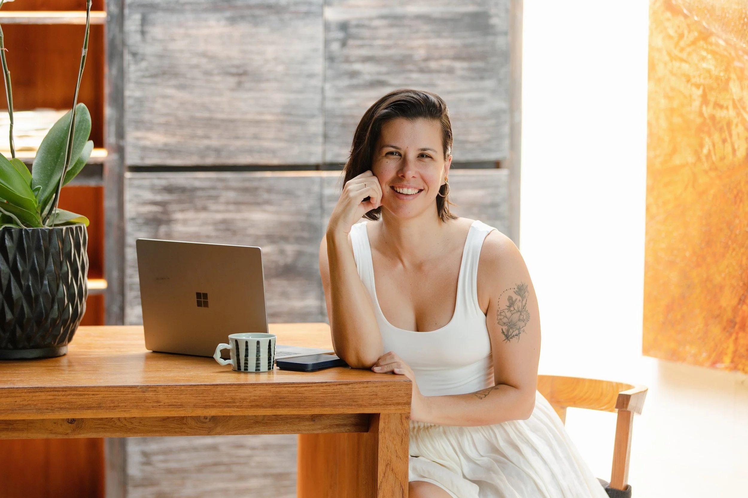 A woman with short dark hair and tattoos on her arm, sitting at a wooden table with a laptop and a mug, smiling and resting her chin on her hand in a cozy, well-lit room with plants and wooden furniture.