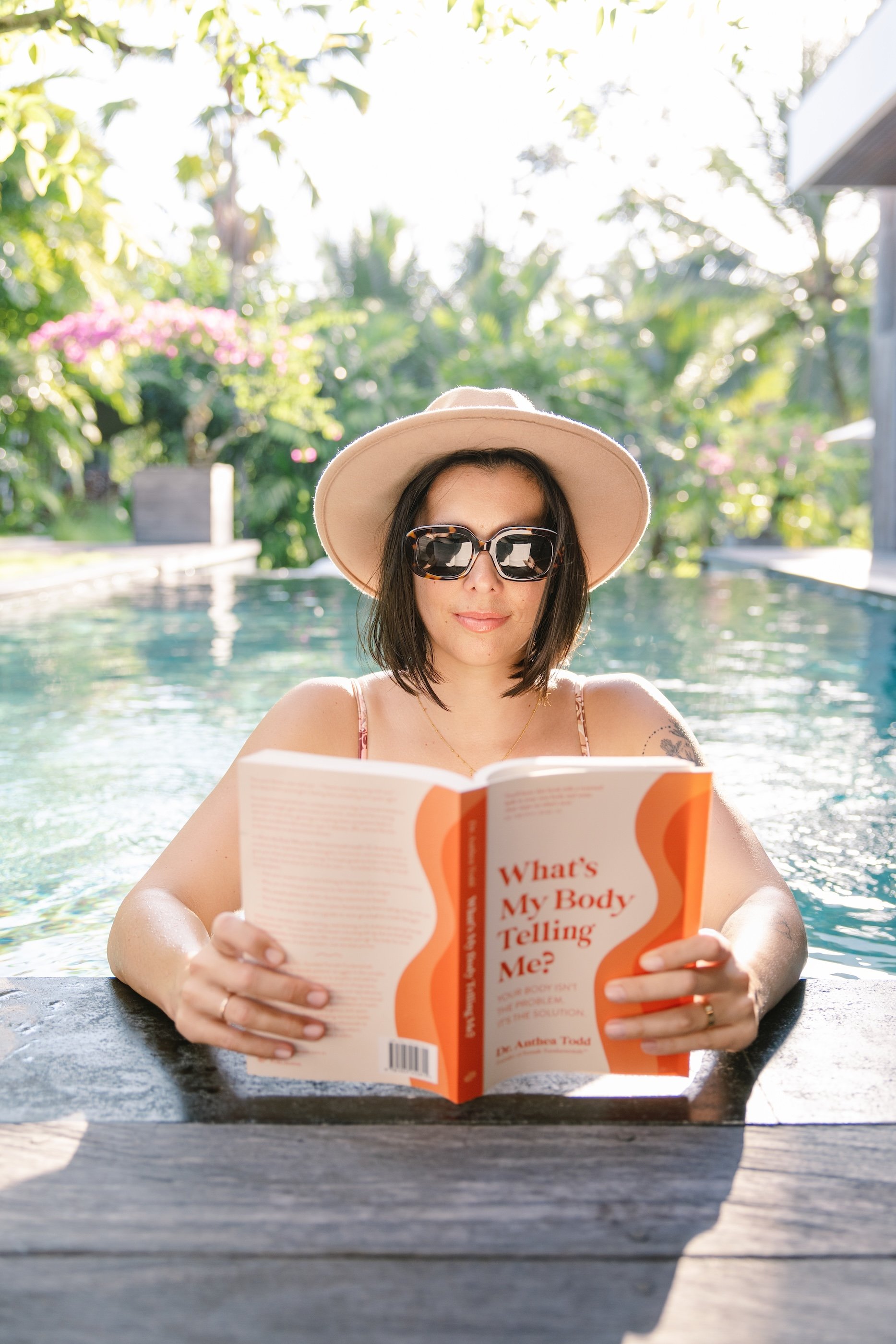 A woman with dark hair wearing sunglasses, a wide-brimmed hat, and a swimsuit, sitting in a swimming pool, reading a book titled 'What's My Body Telling Me?'