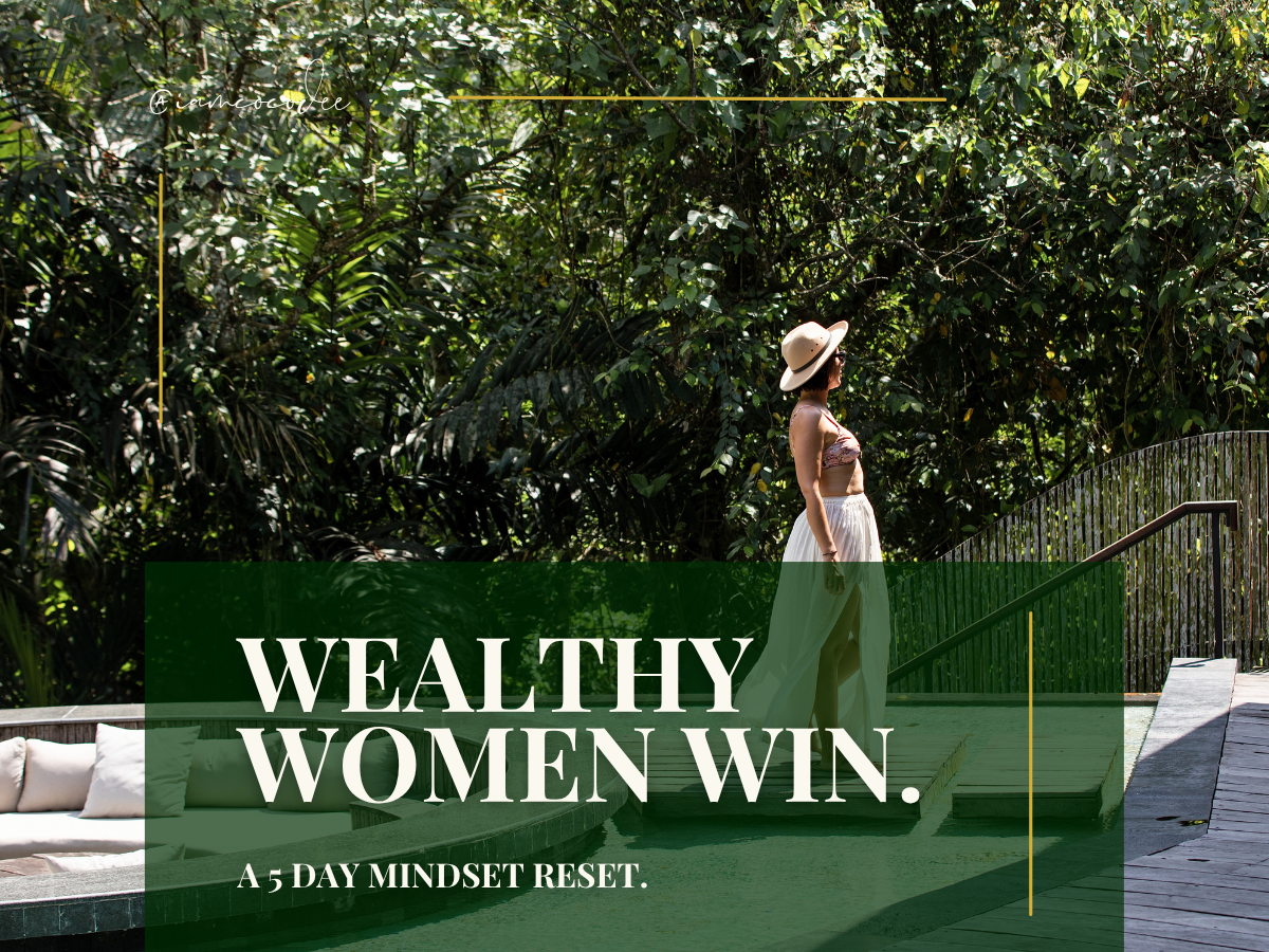A woman in a wide-brimmed hat and summer clothing standing on a wooden deck among lush green foliage, with an overlayed text that reads 'Wealthy Women Win. A 5 Day Mindset Reset.'