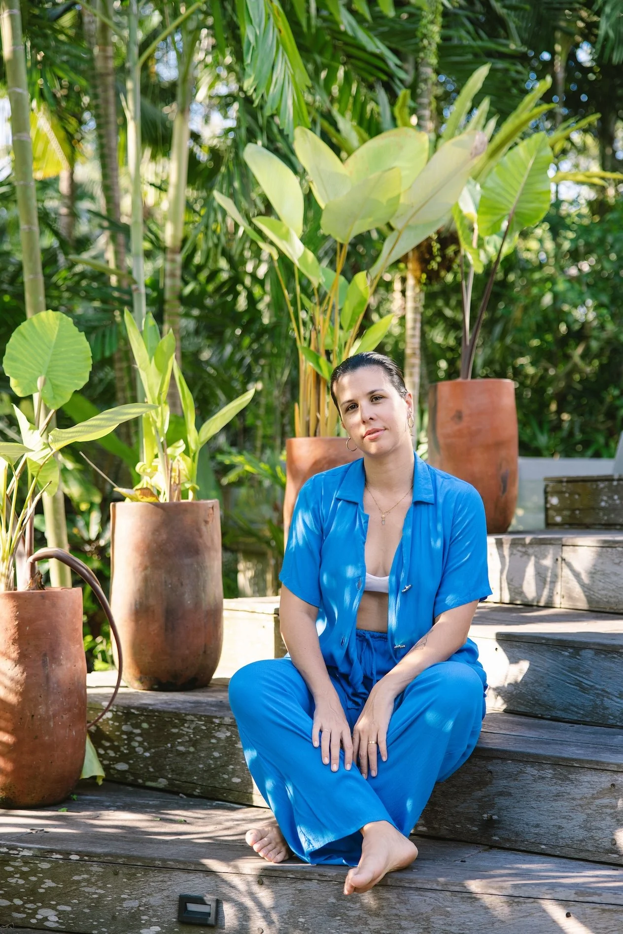 A woman with dark hair in a blue outfit sitting on wooden steps outdoors, surrounded by large potted plants in a lush, green garden.