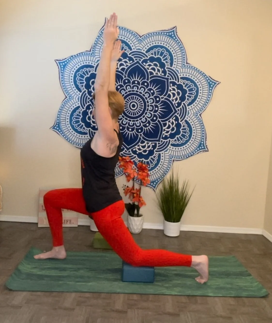 A person doing yoga in a lunge pose with hands raised above the head, standing on a yoga mat with a yoga block under the front leg. The background features a decorative blue mandala wall hanging, a potted plant, and artificial flowers.