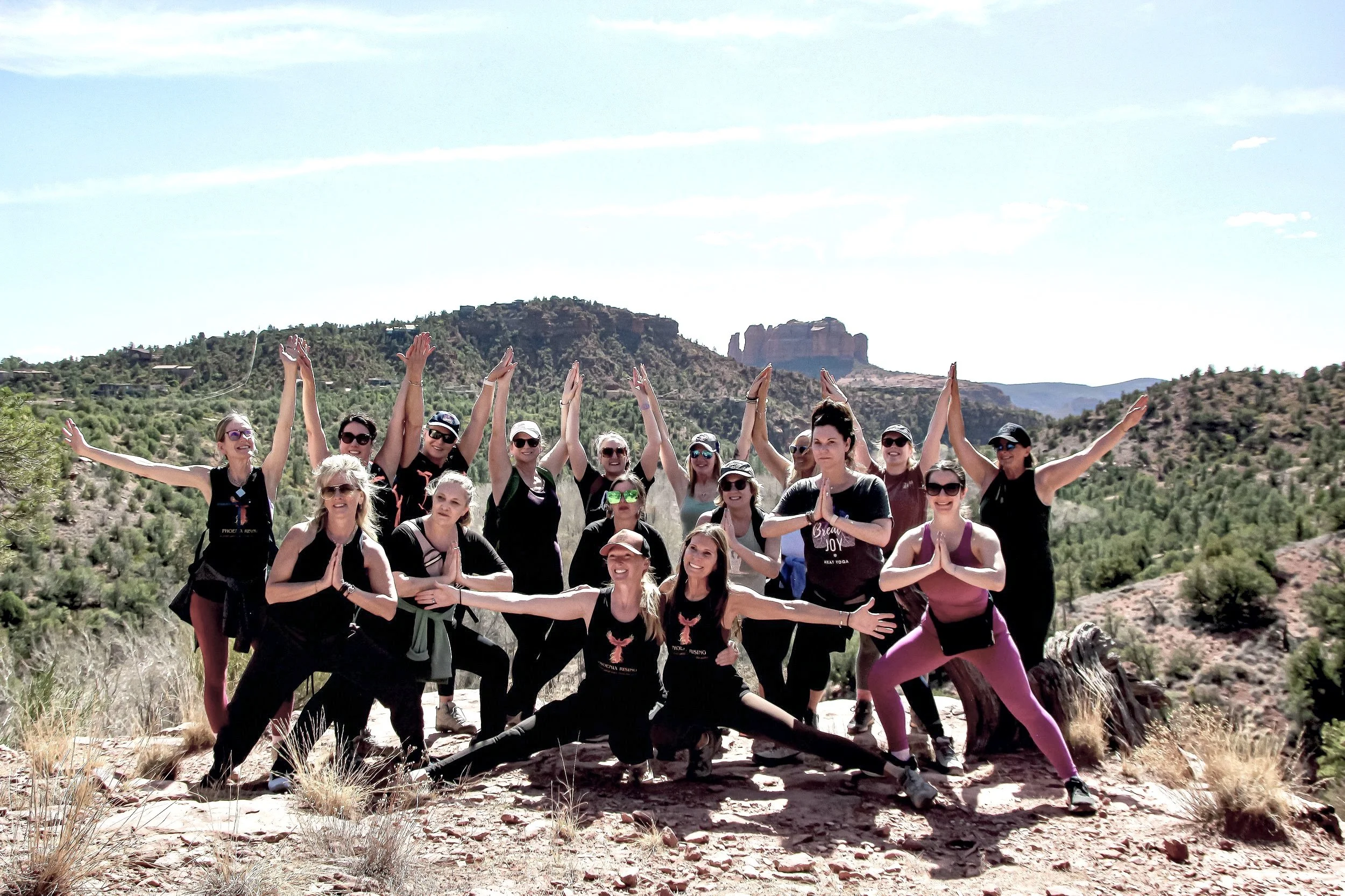 Group of women practicing yoga outdoors on a rocky ledge, surrounded by desert landscape with hills and rock formations in the background.