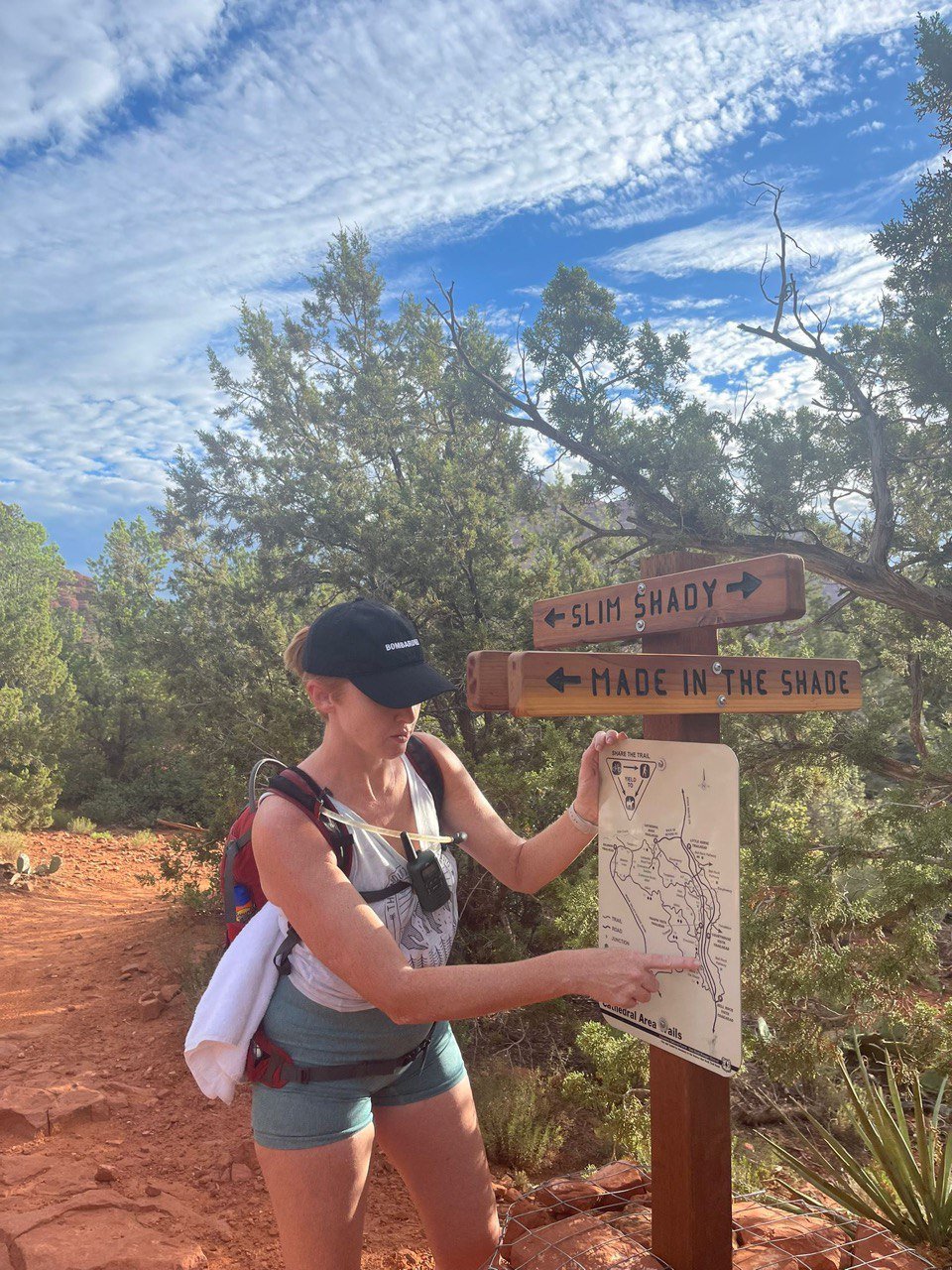 A woman with a backpack and a black cap pointing at a trail map next to a signpost with directions to 'Slim Shady' and 'Made in the Shade' in a desert landscape.
