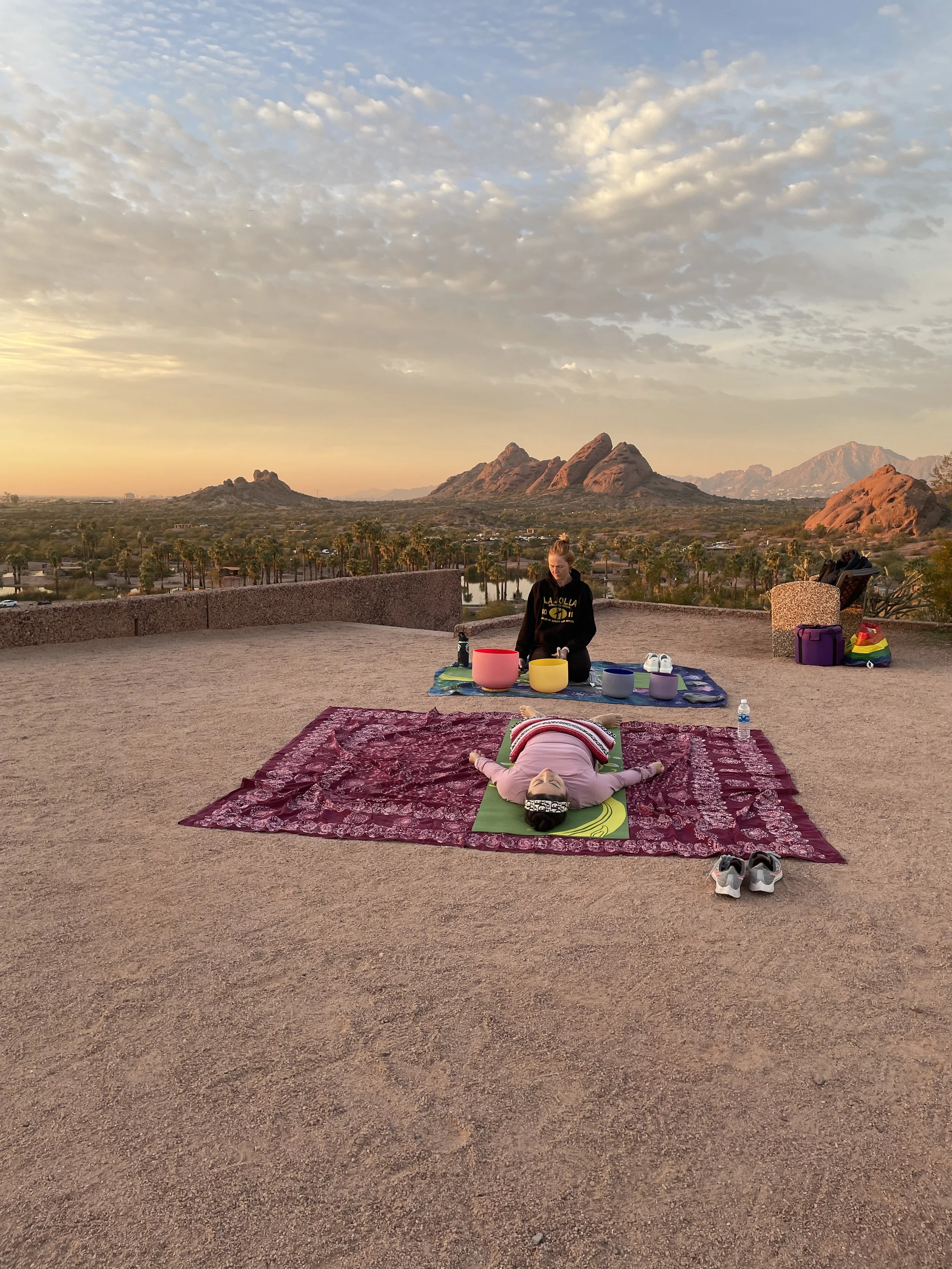 A woman lying on a green yoga mat on a pink rug, with a person sitting behind her playing singing bowls, against a scenic desert landscape at sunset with mountains and clouds in the sky.