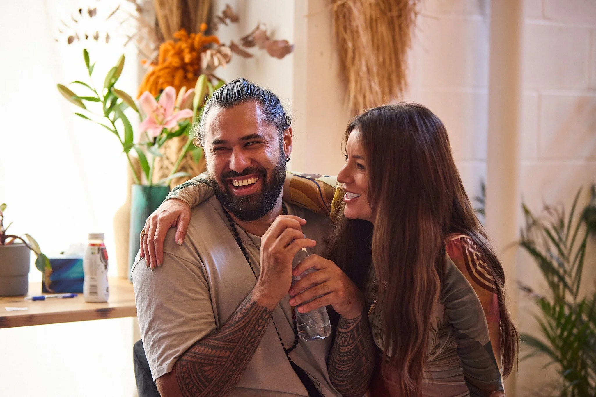 Dr Sarah Jane and Jesse (teacher and practitioner) sharing a joyful moment while teaching. Sarah has long brown hair and Jesse has a beard and tattooed arm, embracing and laughing indoors with plants and flowers in the background.