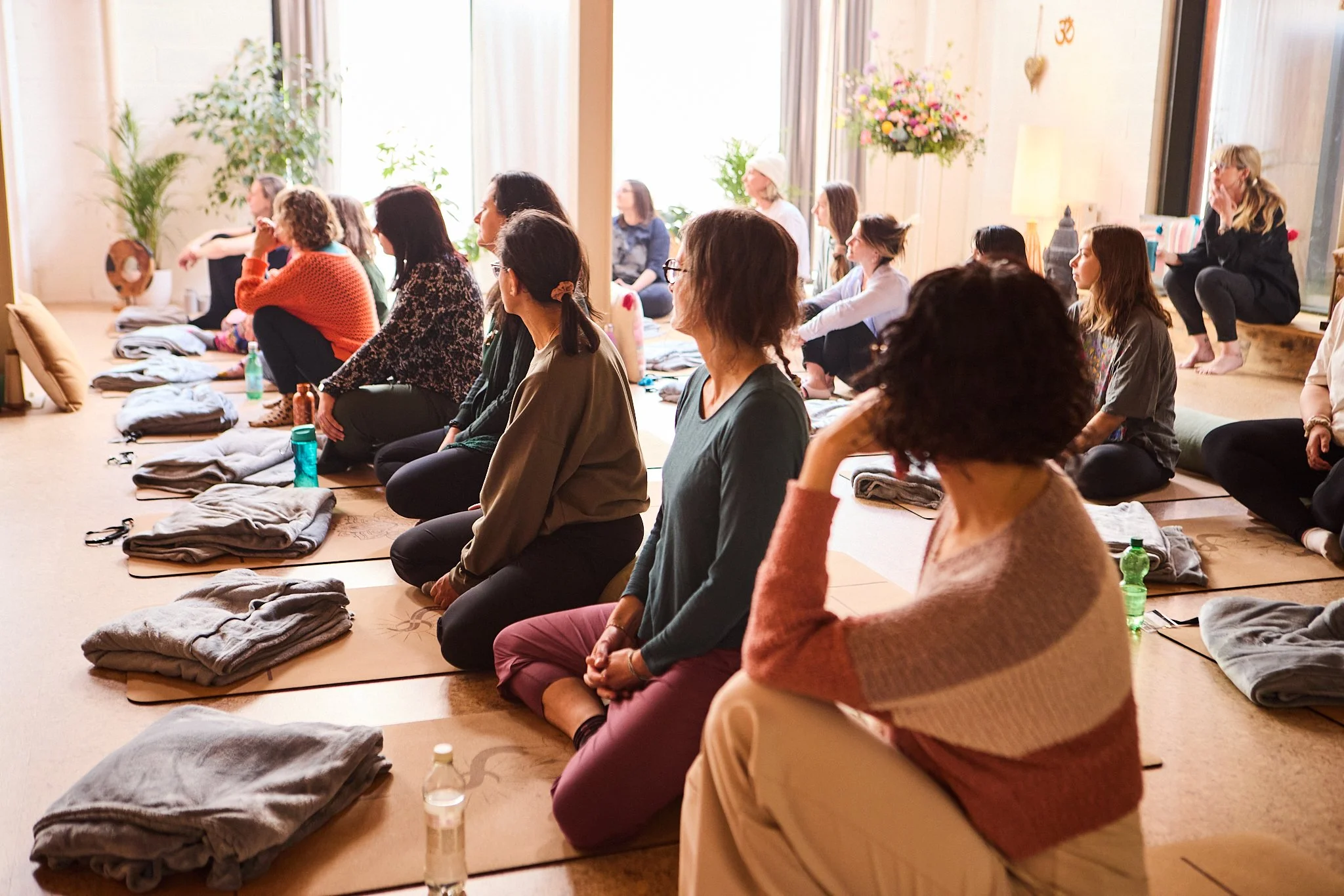 Group of Spinal Energetics students indoors, sitting on yoga mats with folded blankets, facing forward during a session with the teaching team, some sitting cross-legged, in a well-lit room with large windows, plants, and decorative flowers.