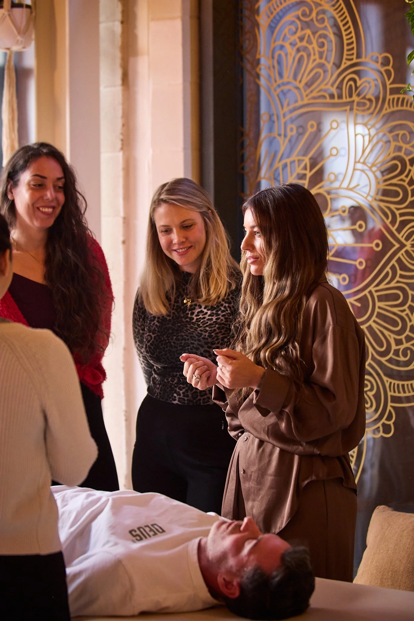Three students are gathered with Dr Sarah around a man lying on a bed, engaged in conversation about Spinal Energetics, with a decorative wall behind them.