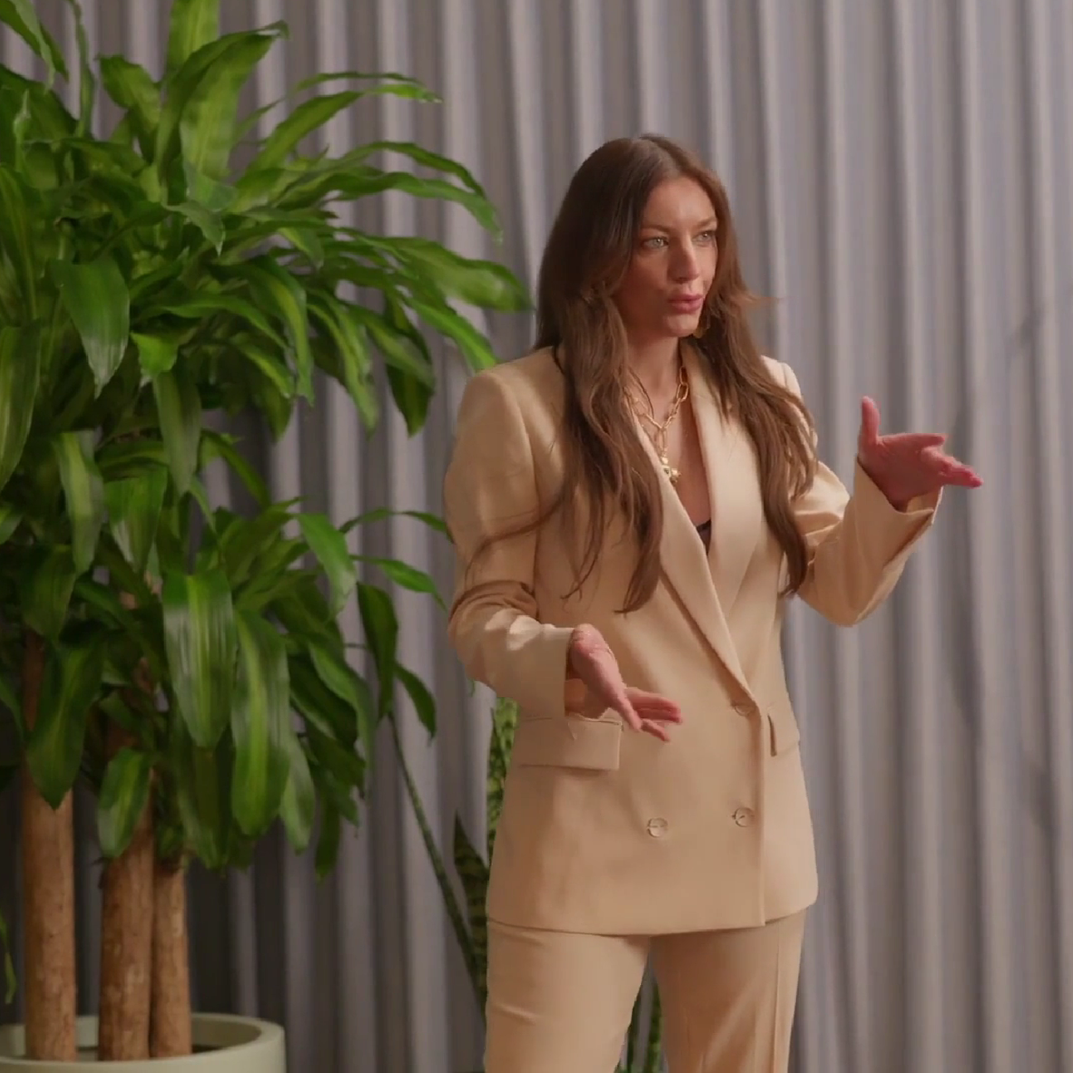 Dr Sarah Jane with long brown hair, wearing a beige suit, is gesturing with her hands while speaking in front of a gray curtain and a large green plant in an Online Clinic video.