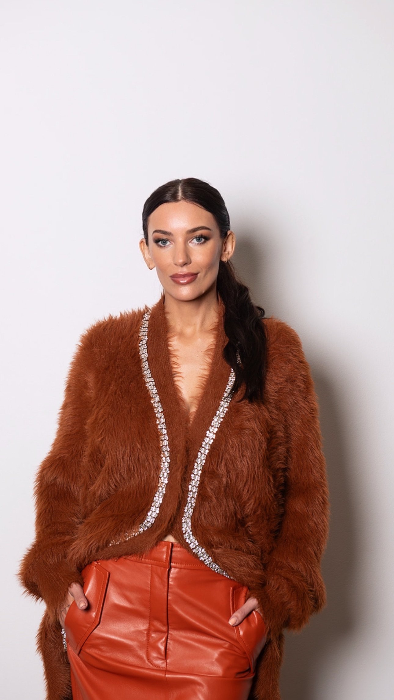 Dr Sarah Jane. She's a woman with dark brown hair styled in a loose wave, wearing a brown faux fur jacket with silver embellishments along the edges, and high-waisted red leather pants, standing against a plain white wall.