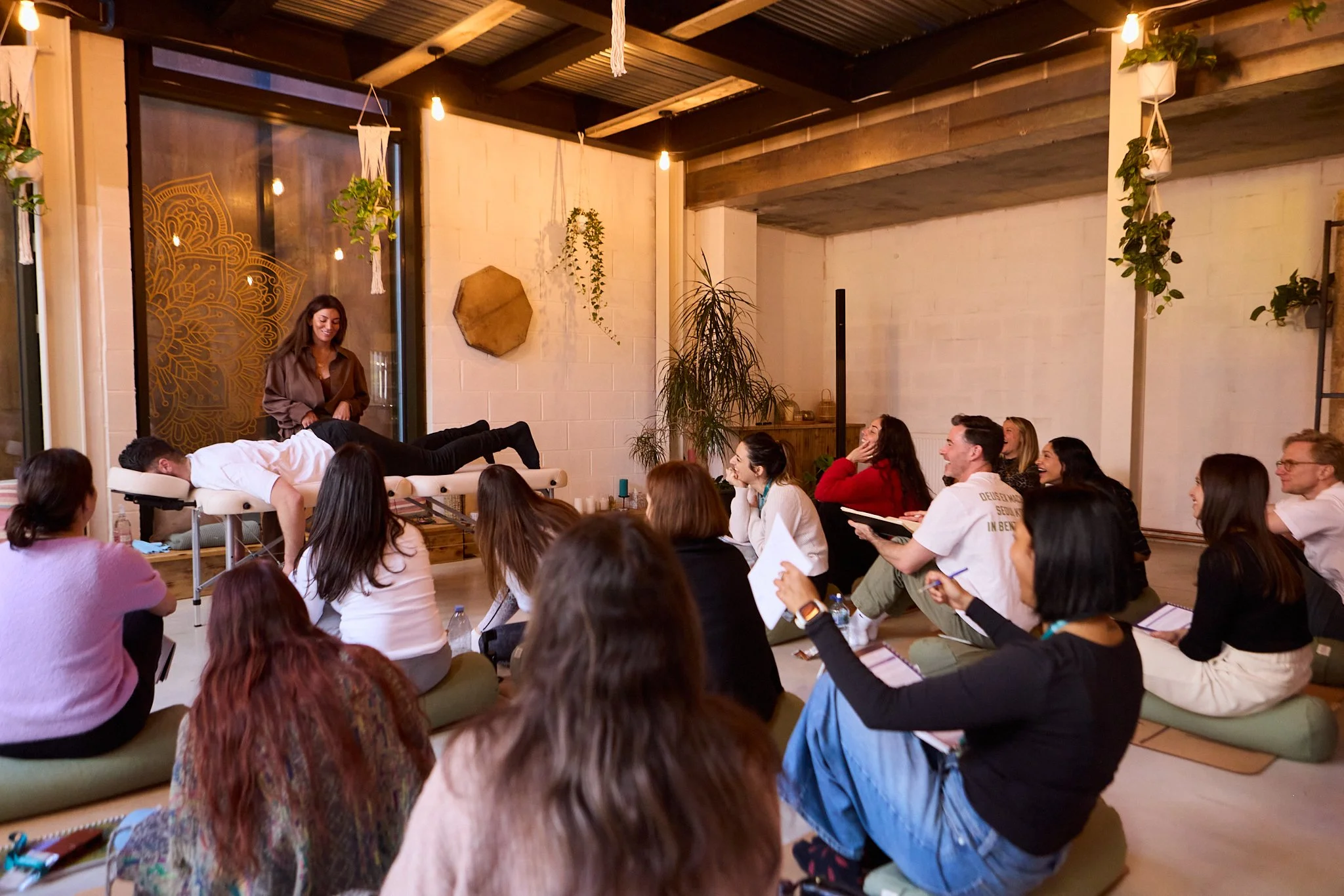 A group of students sitting on cushions on the floor watching a Spinal Energetics demonstration with Dr Sarah Jane and master teacher Pat Chirico during a course, in a cozy, decorated room with plants and warm lighting.