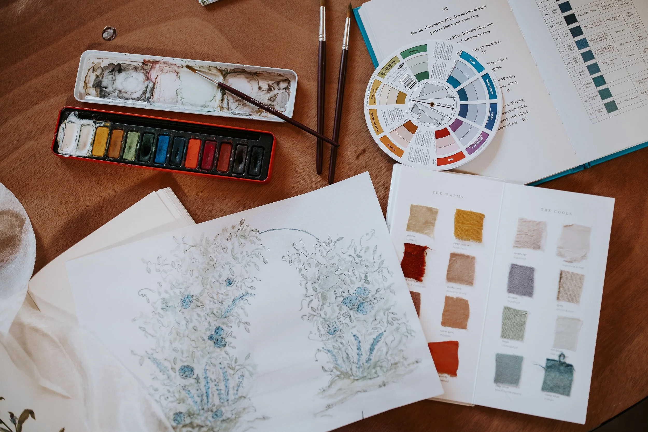 An artist's workspace includes watercolor paints, brushes, a color wheel, a sketch of a floral arrangement, fabric swatches, and open books with color charts.