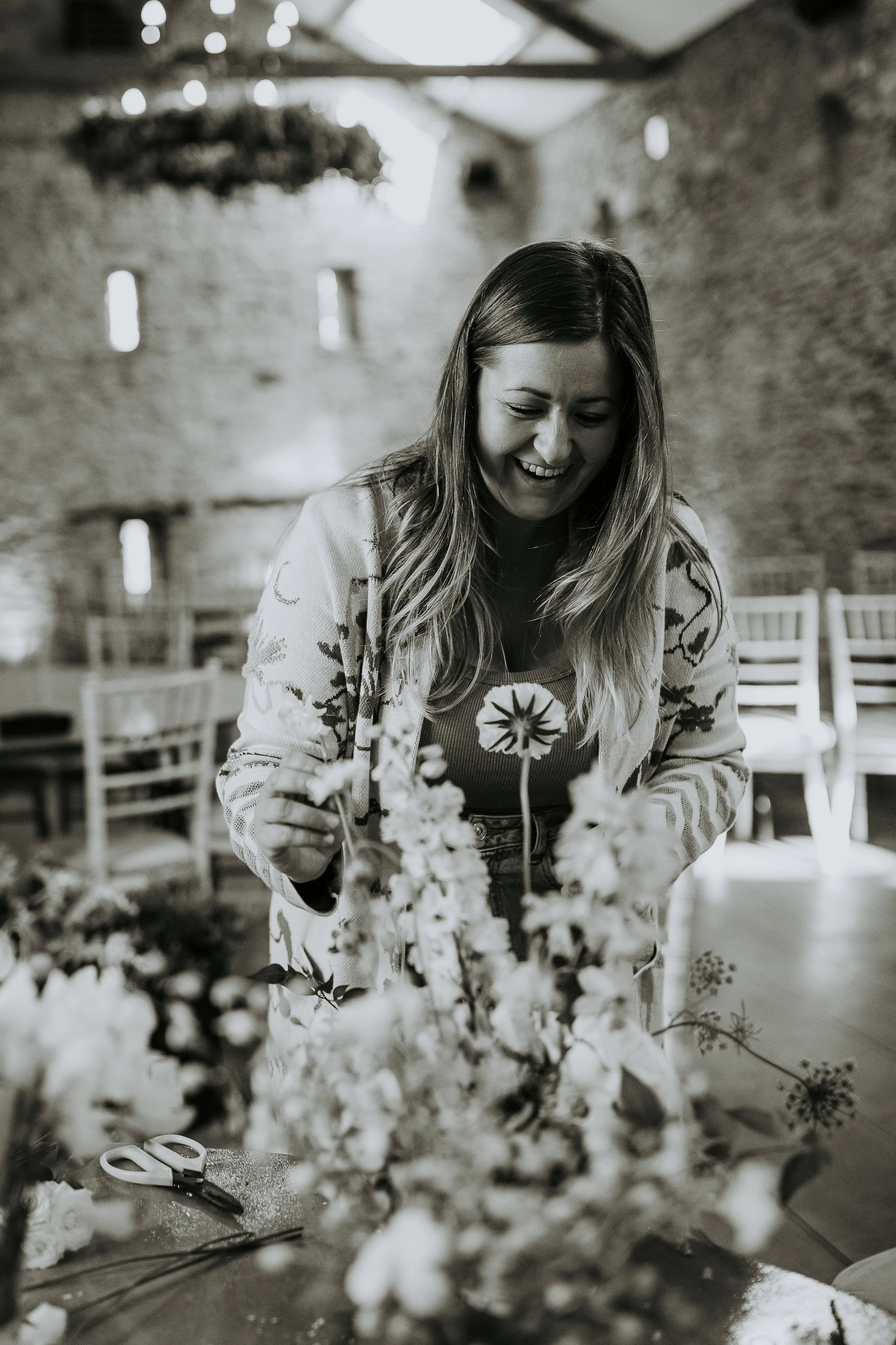 Woman smiling and arranging flowers on a table in a rustic indoor setting.