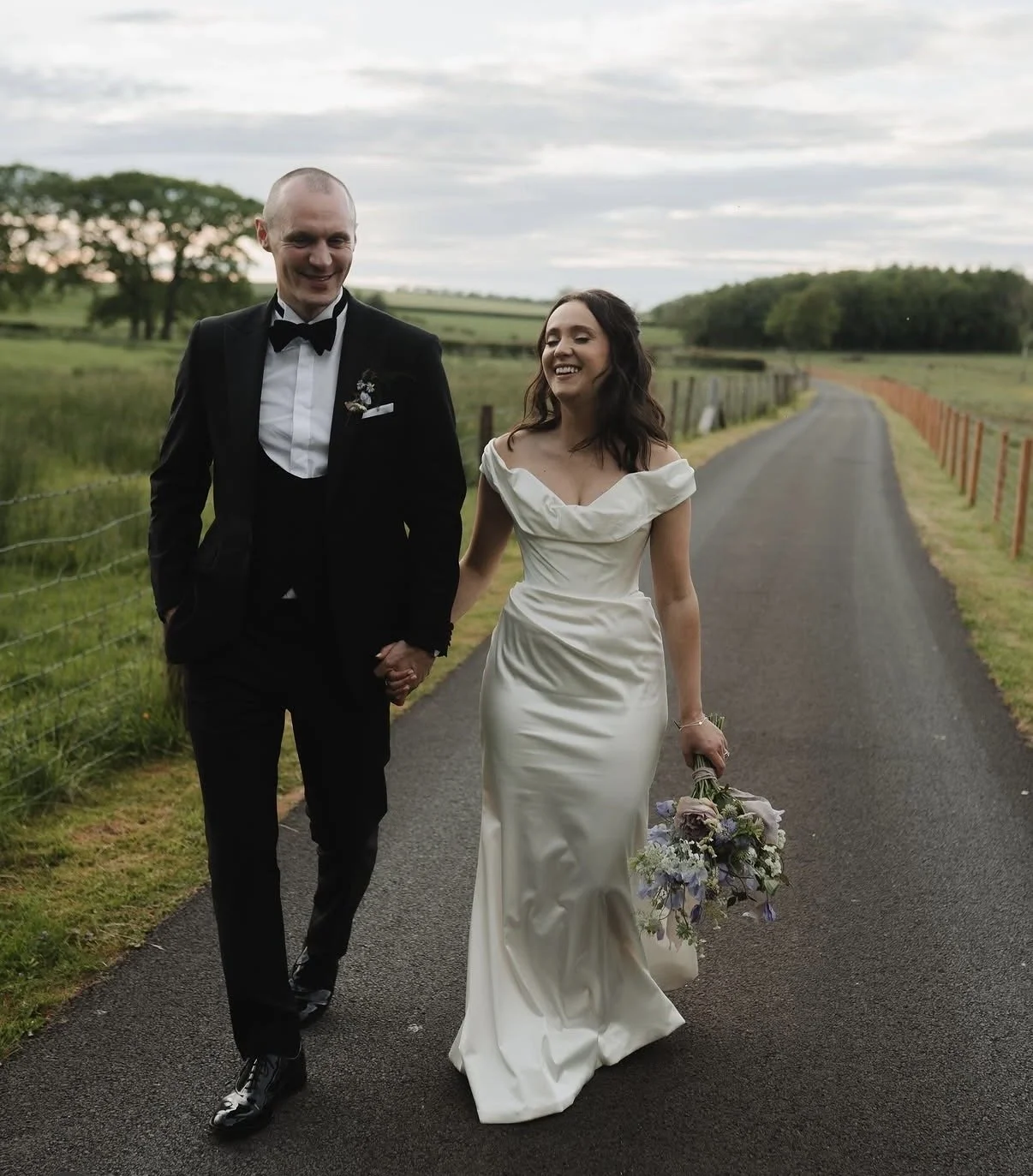 A bride and groom dressed in wedding attire and holding her bouquet, walking hand in hand on a country road, with a grassy field and trees in the background, and cloudy sky overhead.