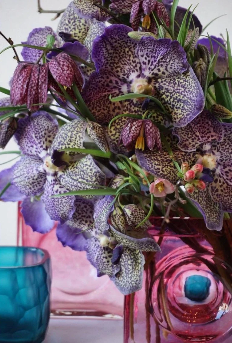 Un bouquet de fleurs avec des orchidées violettes et blanches dans un vase rose en verre, avec un verre bleu à côté.