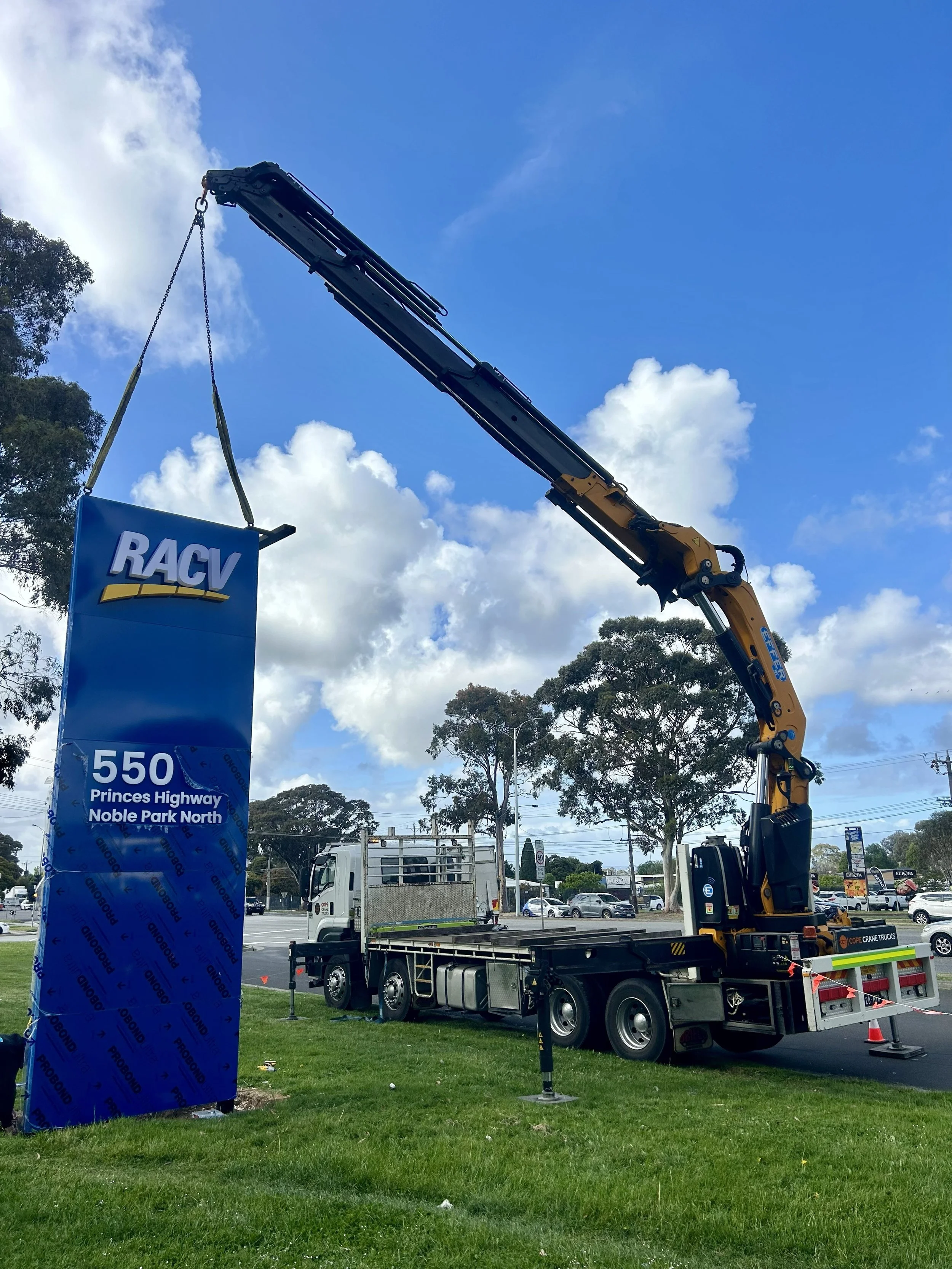 Cope Crane Trucks' crane truck positioned on the side of the road with the Effer truck-mounted crane crane extended and lowering a RACV sign in place. The image is used as an example of Cope Crane Truck's crane truck hire services.
