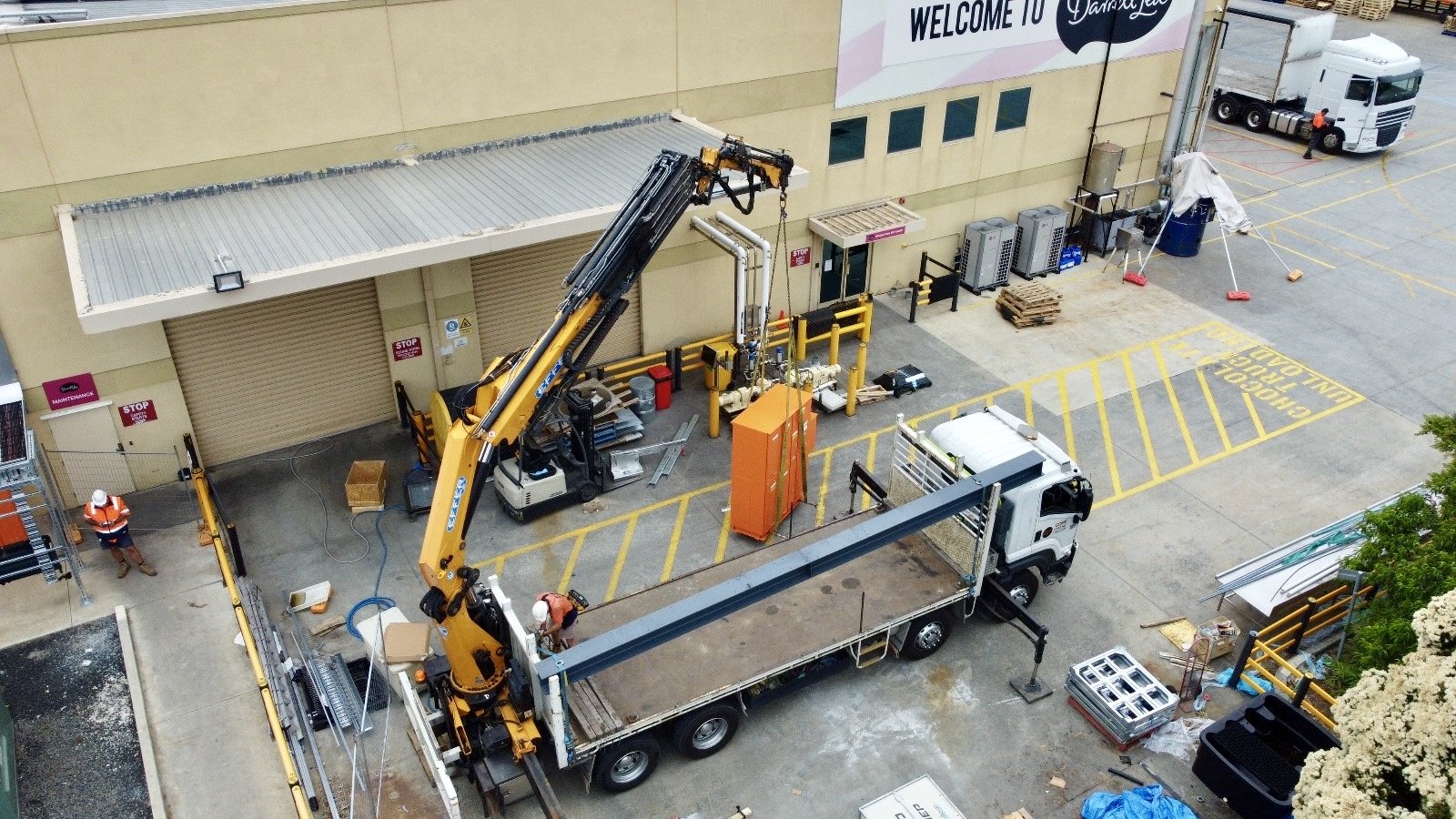 Angled bird's eye view action shot of Cope Crane Trucks crane truck hire for Darrell Lea in Melbourne. Image includes an Effer truck mounted crane moving a large orange piece of machinery onsite.