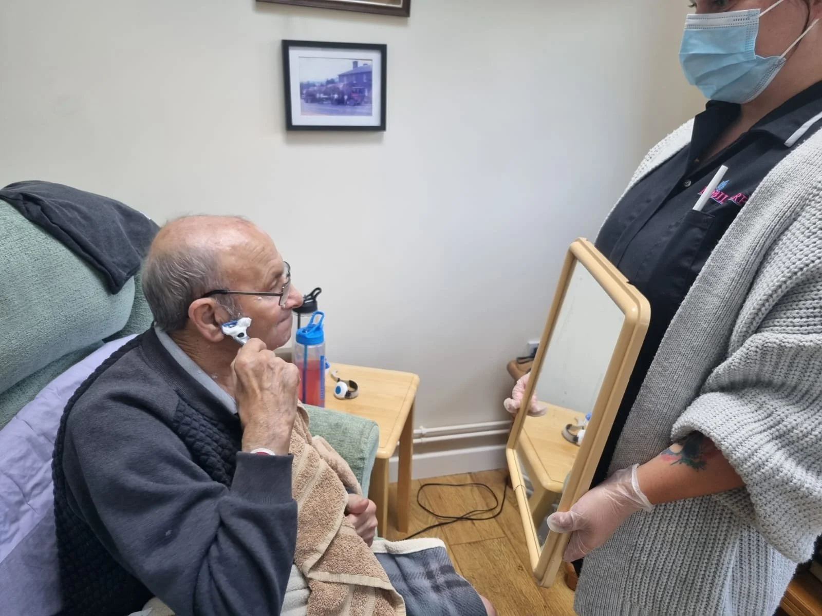 Older man shaving with a nurse's assistance.