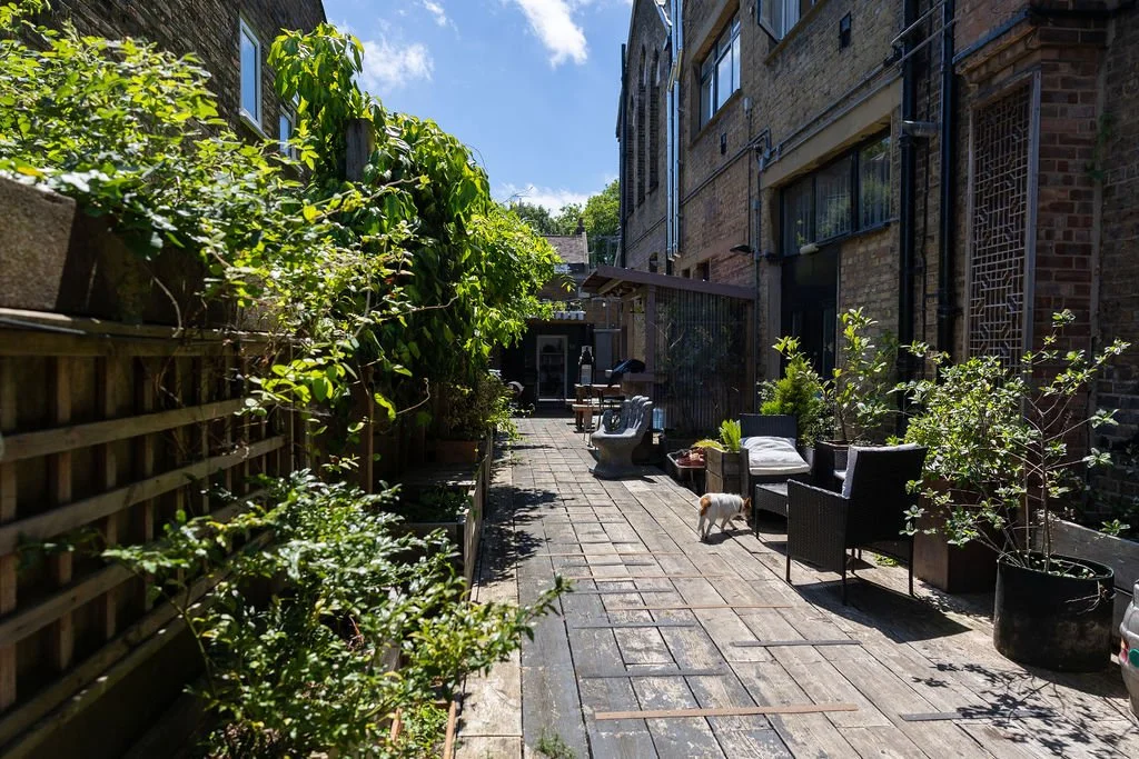 Outdoor urban patio or alleyway with potted plants, outdoor furniture, and a small dog, surrounded by brick and wooden buildings and a wooden fence on the left, under a partly cloudy sky.